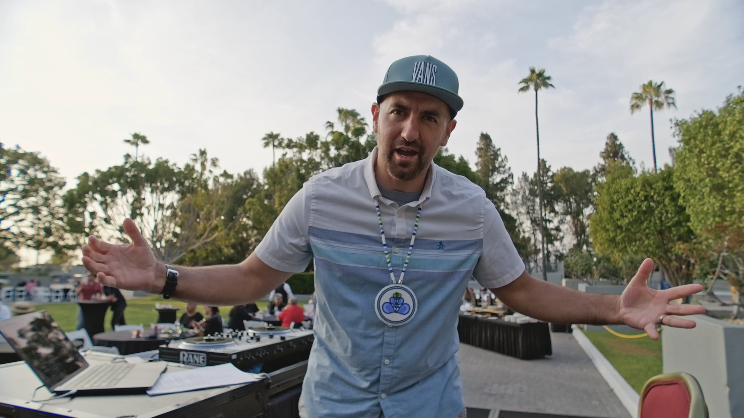 A man with a beard and wearing a blue cap and white shirt with blue stripes is gesturing with his arms open at an outdoor event. There is a DJ setup with headphones and a laptop on a table nearby, and people are sitting at tables in the background. T
