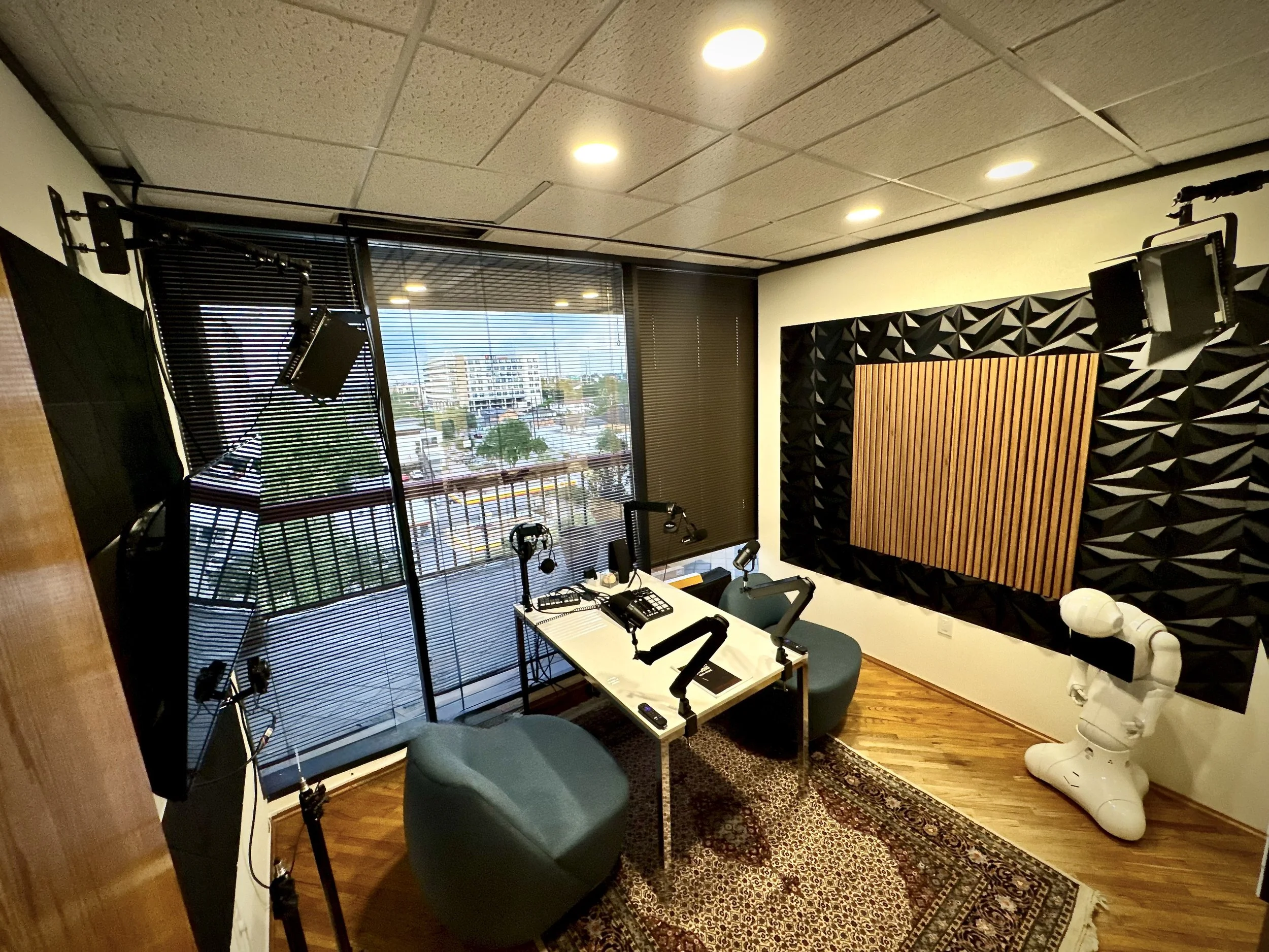 A podcast studio with two chairs, a white table with microphones, headphones, and a control panel, enclosed by soundproofing panels on the walls. There are large windows with blinds overlooking a cityscape.