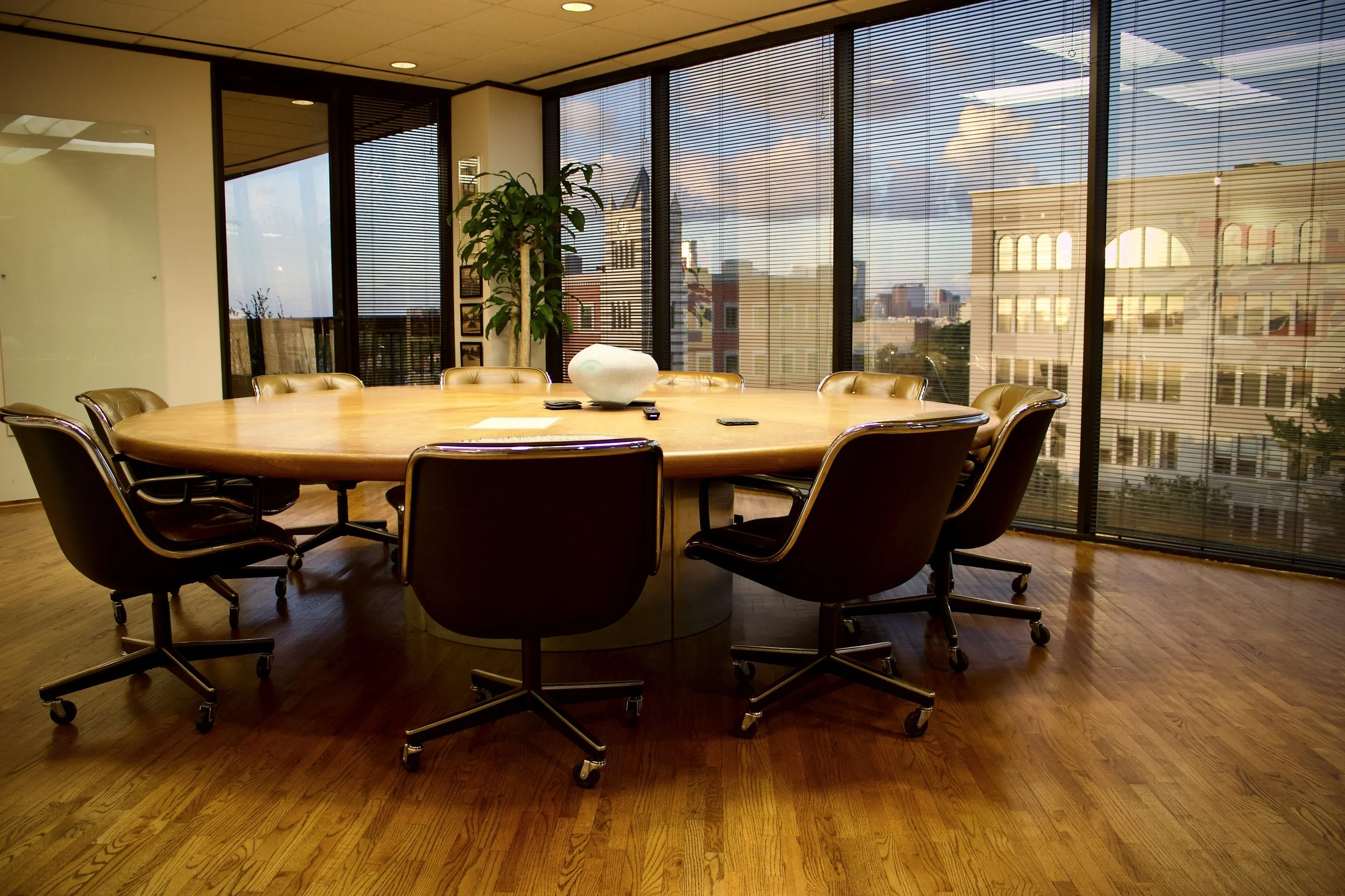 Conference room with large wooden table surrounded by black office chairs, large windows with blinds, and cityscape view outside.