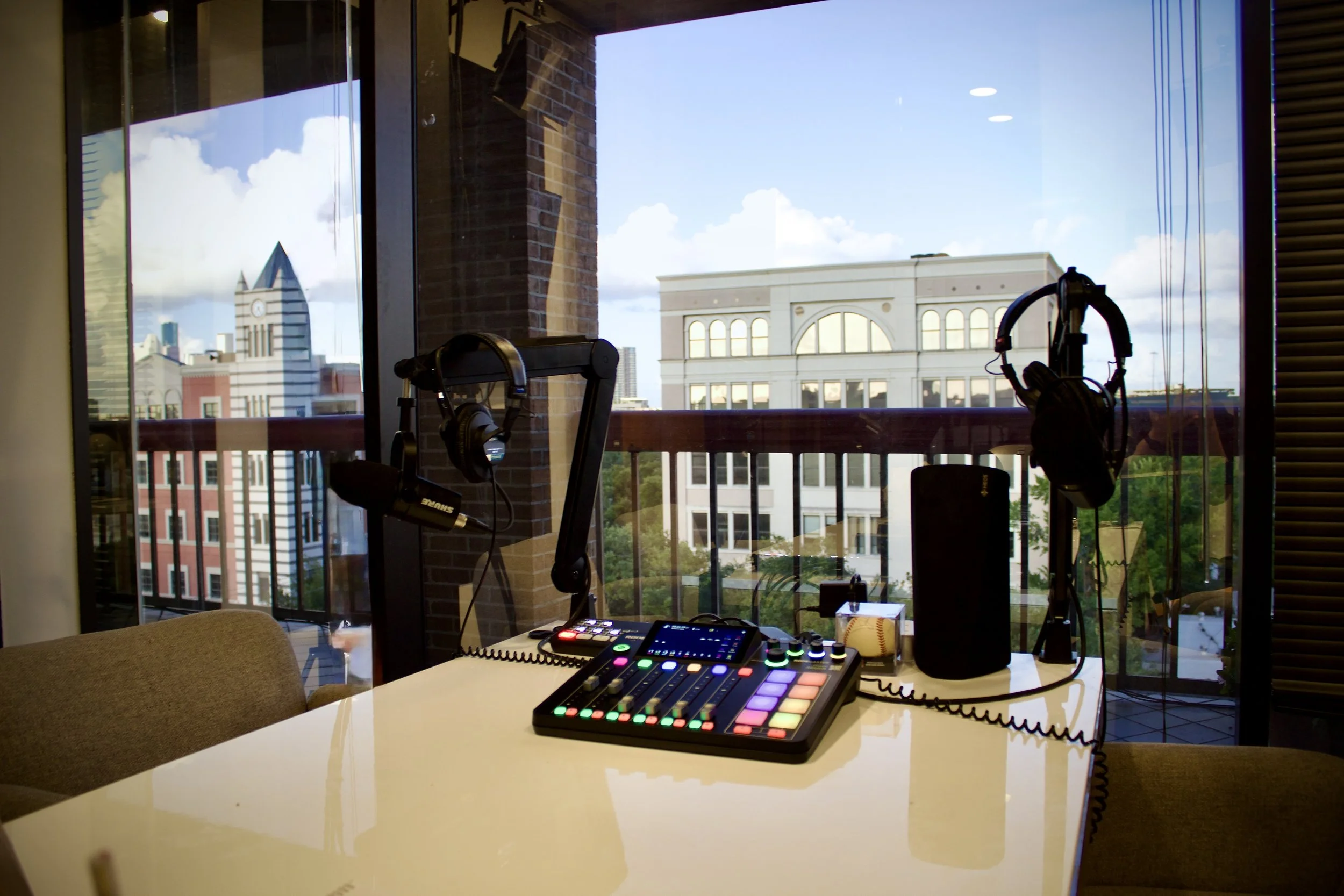 DJ equipment setup on a white table inside a room with large windows showing city buildings and a cloudy sky.