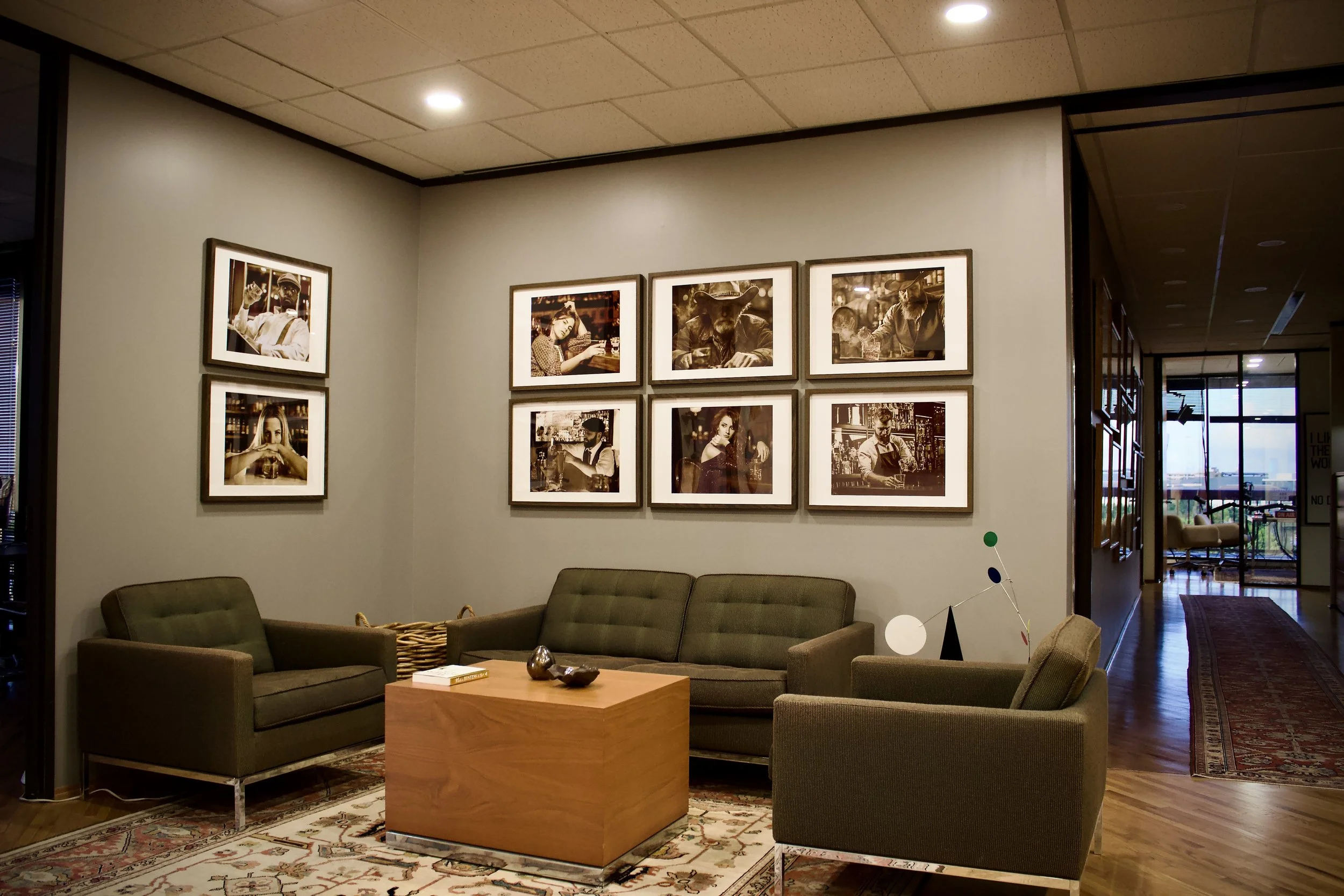 Modern lobby with three green sofas, a wooden coffee table, framed black-and-white photographs on the gray wall, a patterned area rug, and a view of the outdoor area through glass doors.