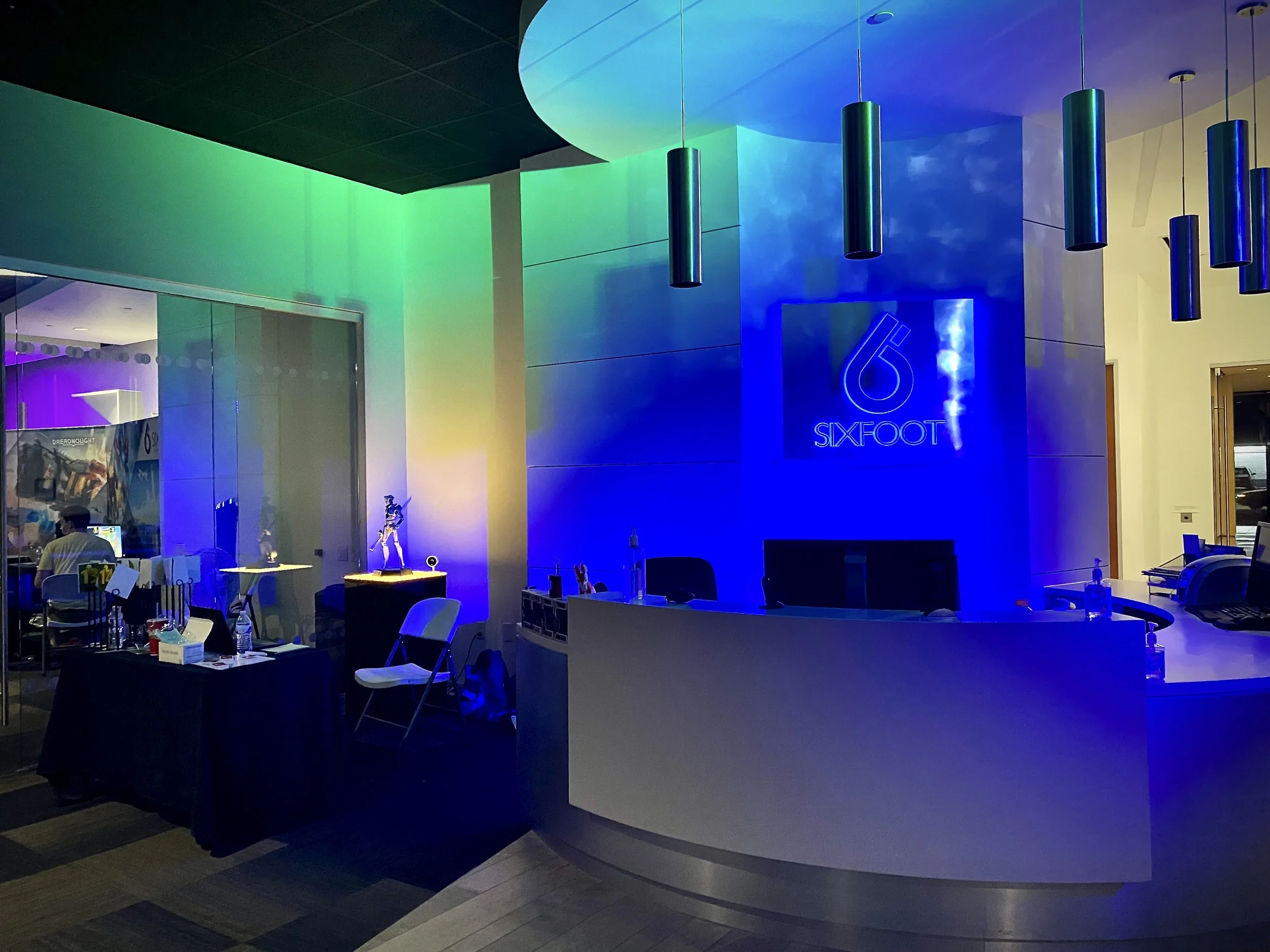 Reception area with colorful rainbow lighting, a desk with a computer, and a neon sign reading '6 SIXFOOT' on the wall.