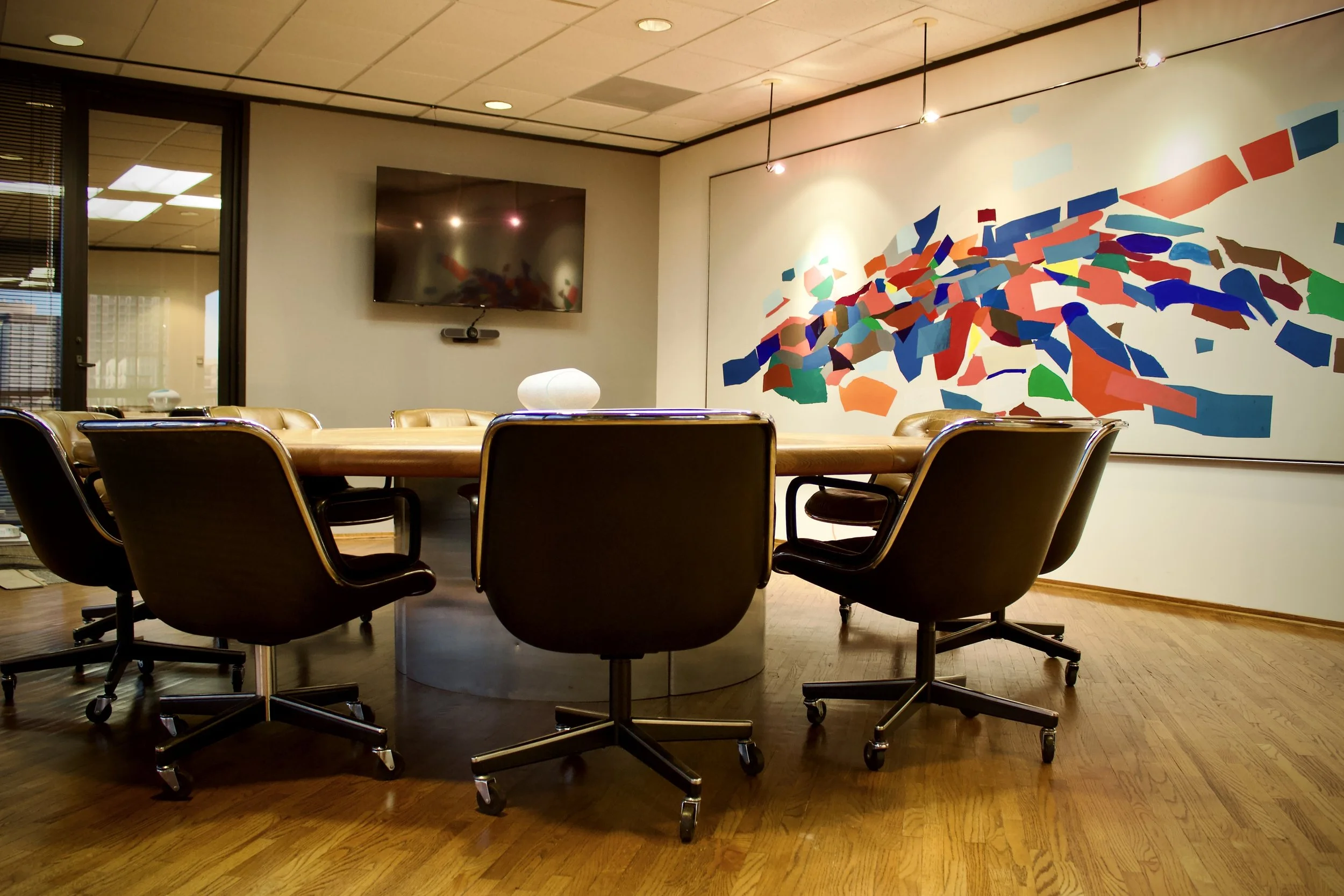 Empty conference room with a large wooden table, black office chairs, a flat-screen TV on the wall, and an abstract colorful mural on the wall.