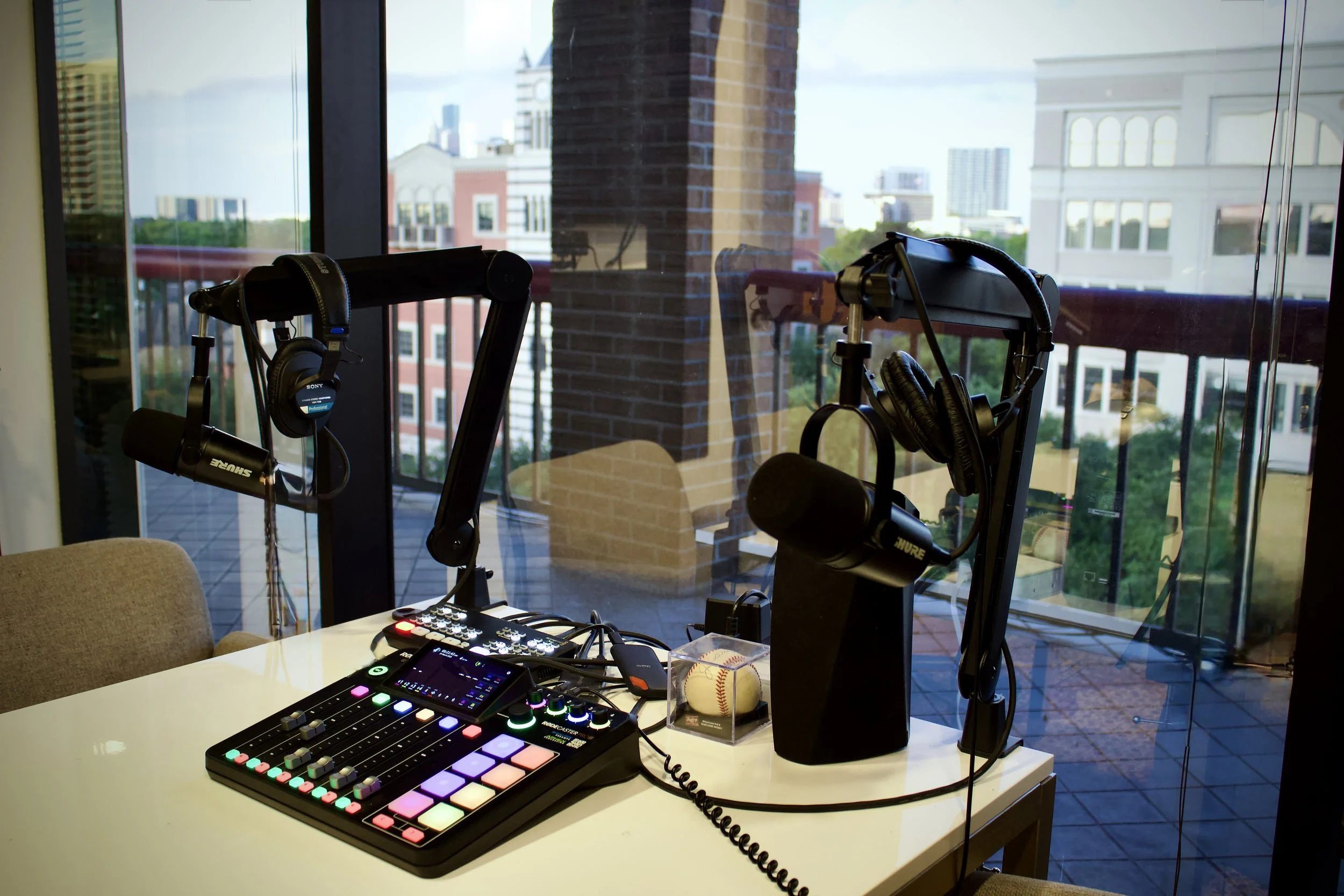 Podcast recording setup with microphones, headphones, mixer on a white table near large windows showing city buildings