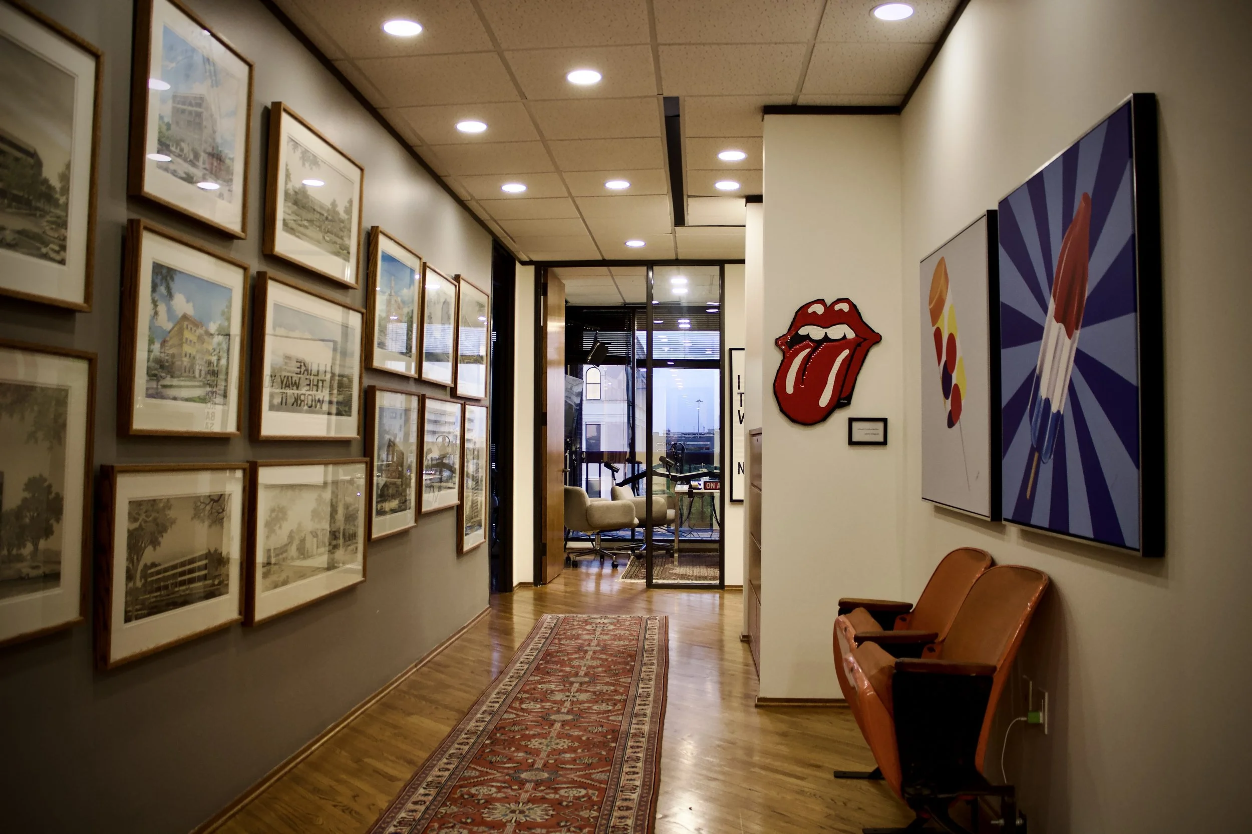 Interior of an art gallery or office space with framed pictures on the left wall and pop art on the right, including the Rolling Stones tongue logo, a lipstick illustration, and a colorful paintbrush. Wooden floor with a patterned rug and a view to t
