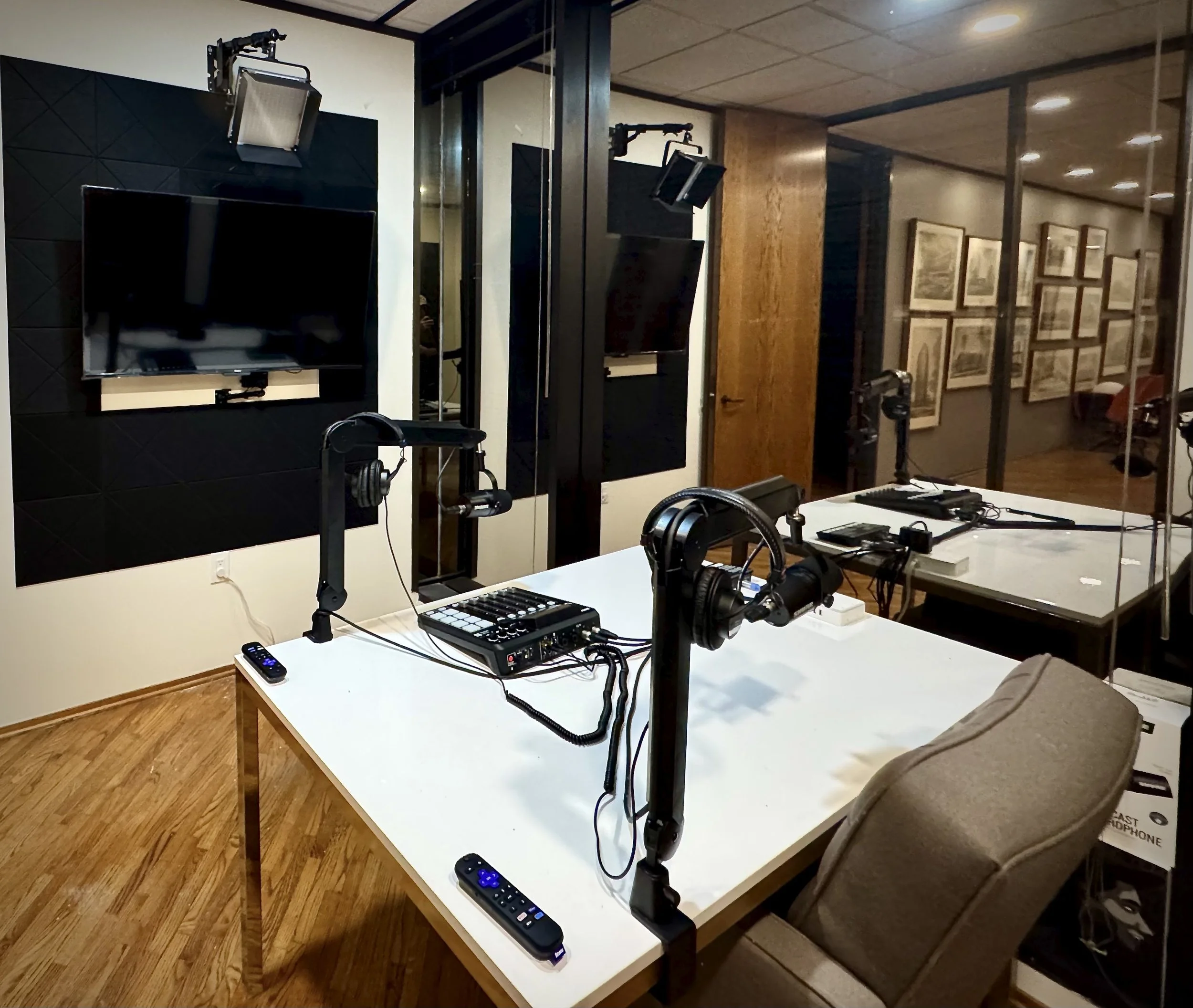 Podcast recording studio with a white table, microphones, headphones, and remote controls, located inside a glass-enclosed room with framed artwork on the walls.