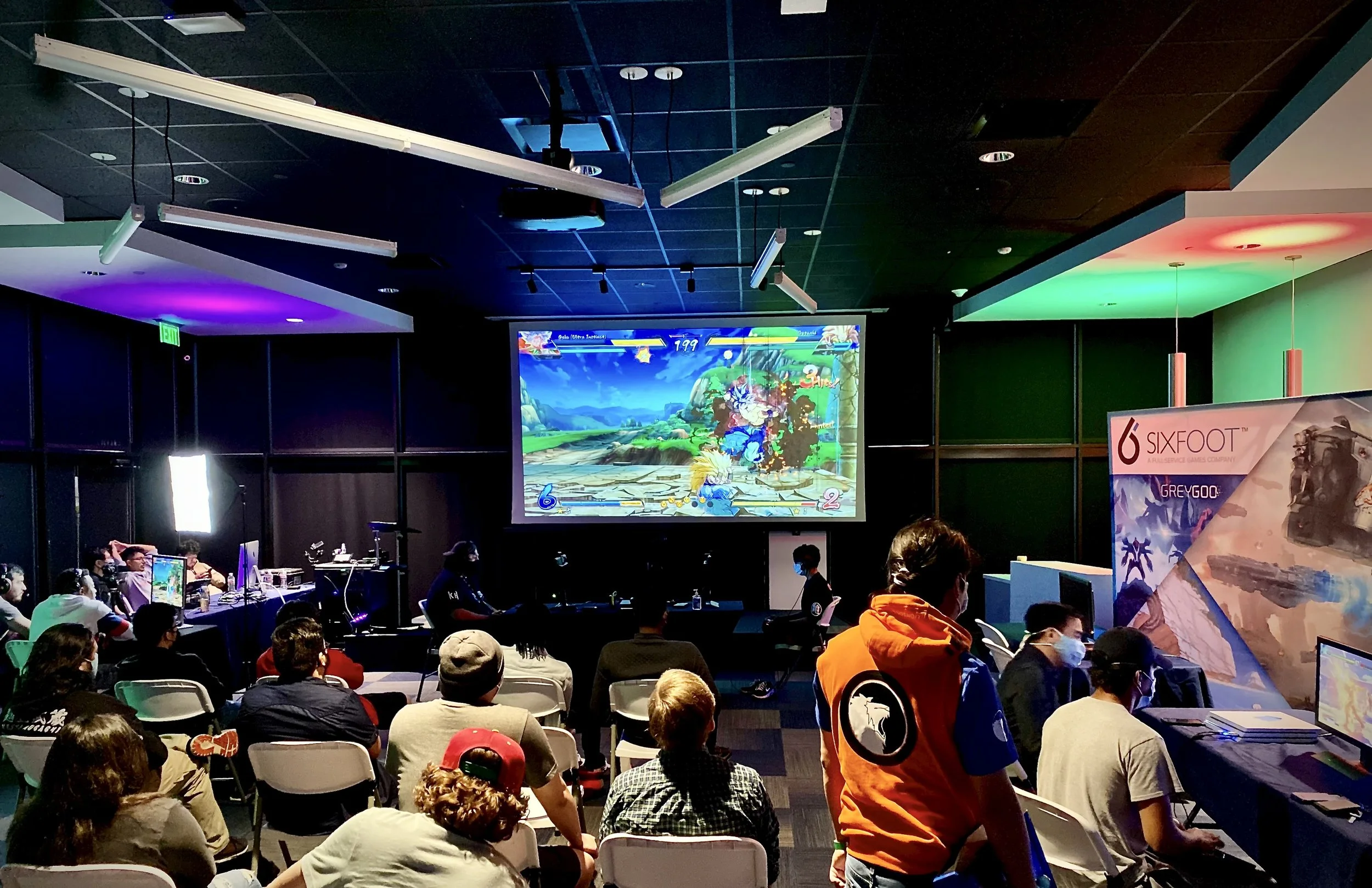 Group of people watching a gaming tournament in a dark room with large screen displaying a fighting game, with colorful lights and gaming equipment on the sides.