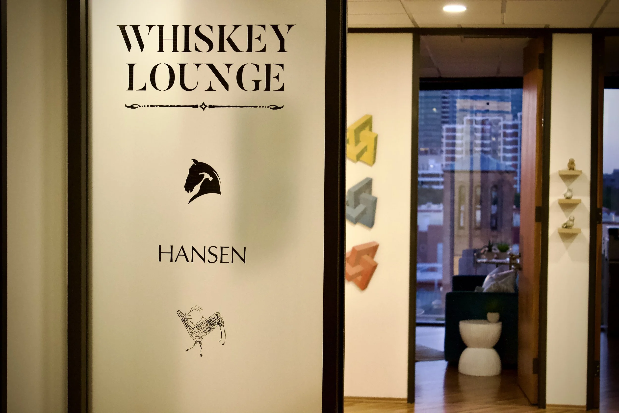 Sign reading 'Whiskey Lounge' with the Hansen logo and a graphic of a horse's head, next to an interior view of a modern lounge area with a cityscape view through large windows.