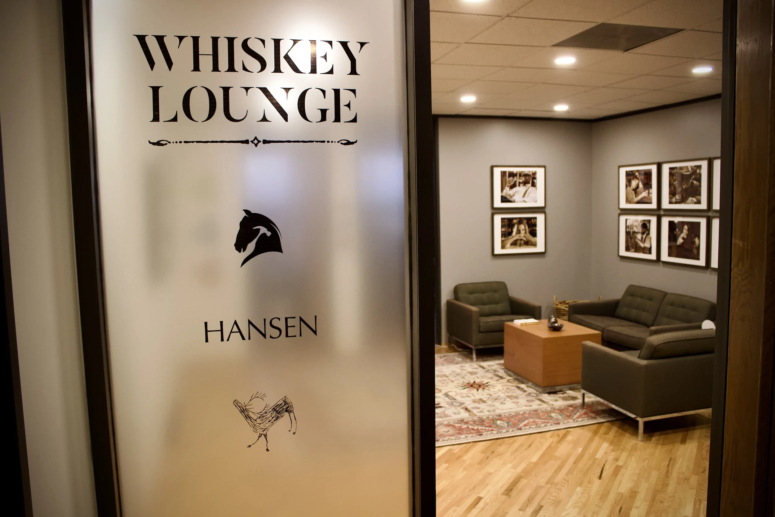 Entrance to Whiskey Lounge with frosted glass door, black text, and a horse logo, revealing a cozy seating area with three gray couches, framed photographs on the wall, and a wooden coffee table inside.