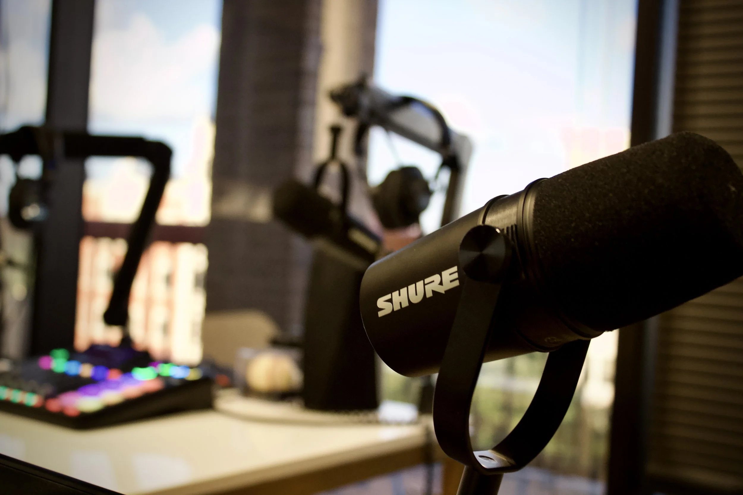 Close-up of a Shure microphone in front of a music production setup, including a keyboard with colorful LED lights, in a room with large windows and a brick wall.