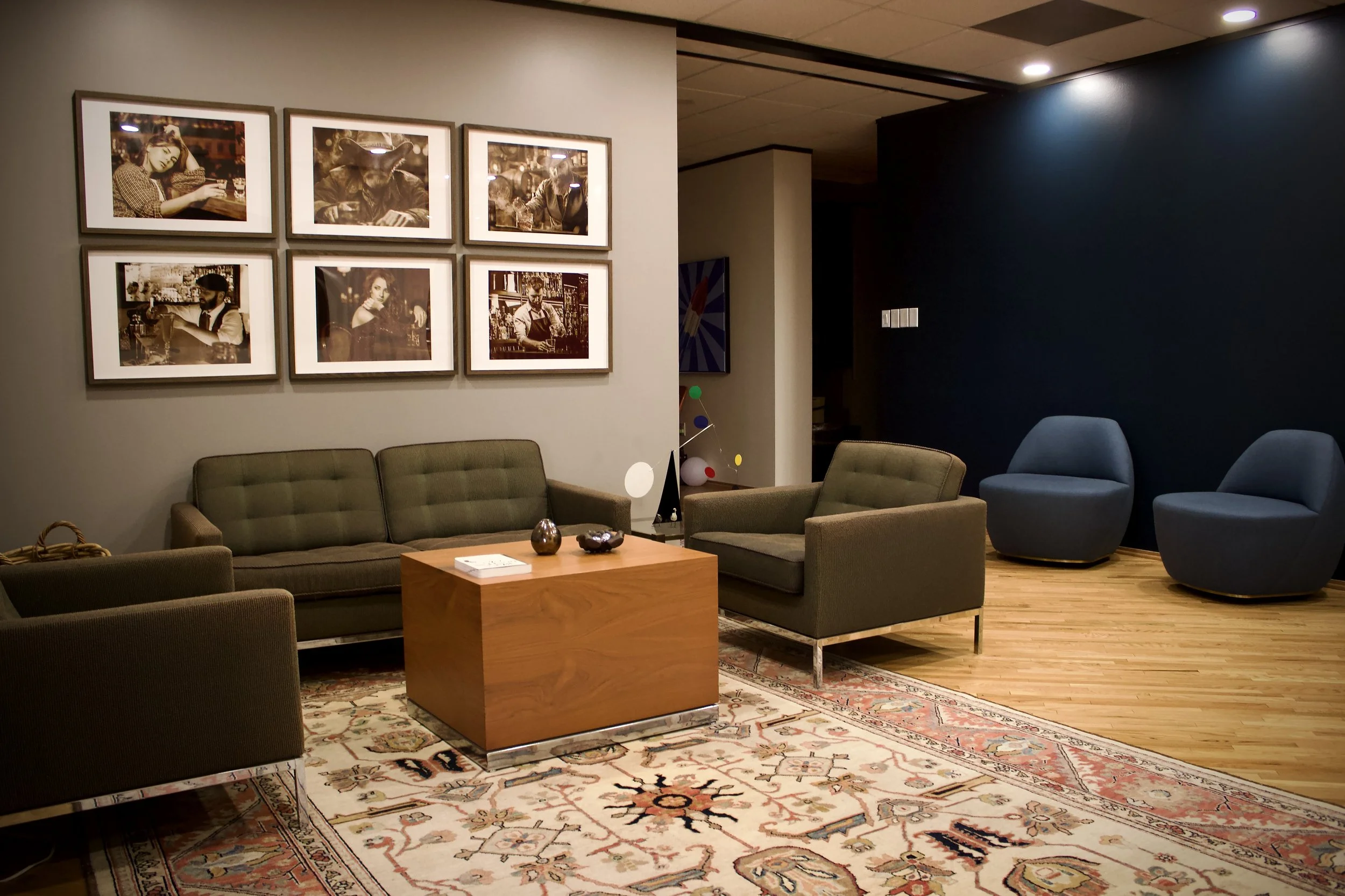 A cozy sitting area with a green sofa and an armchair, a wooden coffee table, framed photographs on the wall, and two modern blue chairs in the background, with a beige and red patterned rug on hardwood floors.