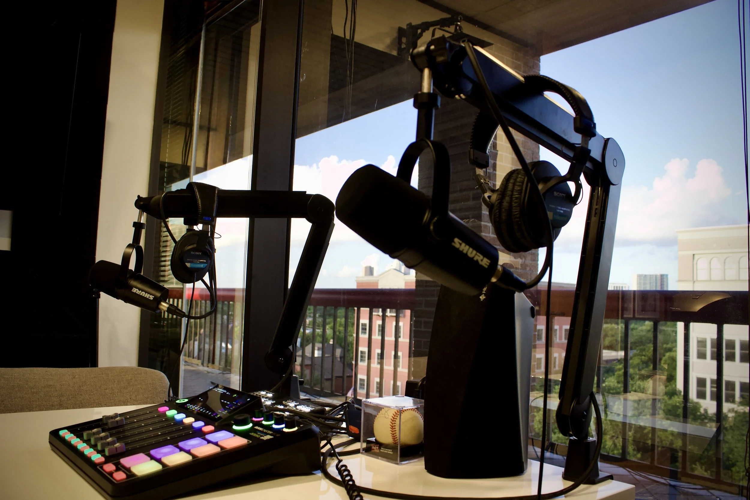 A podcast recording setup on a table with a colorful digital audio mixer, two microphones, two pairs of headphones, and a baseball resting in a clear case, with a background of a balcony and city buildings outside large glass windows.