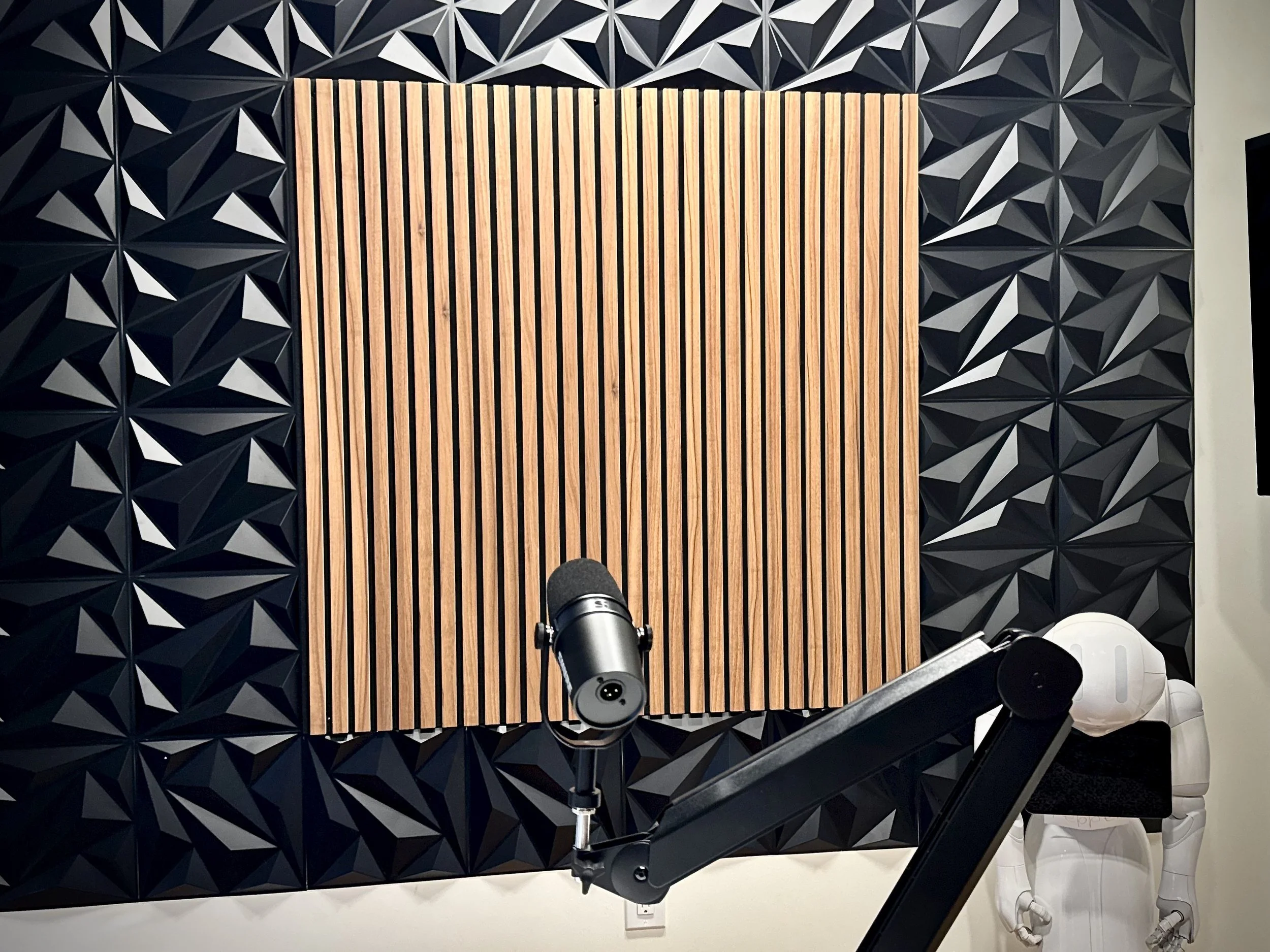 Recording studio with black acoustic foam panels and wooden soundproofing panels, a microphone on a stand, and a white humanoid robot with a black plush seat.