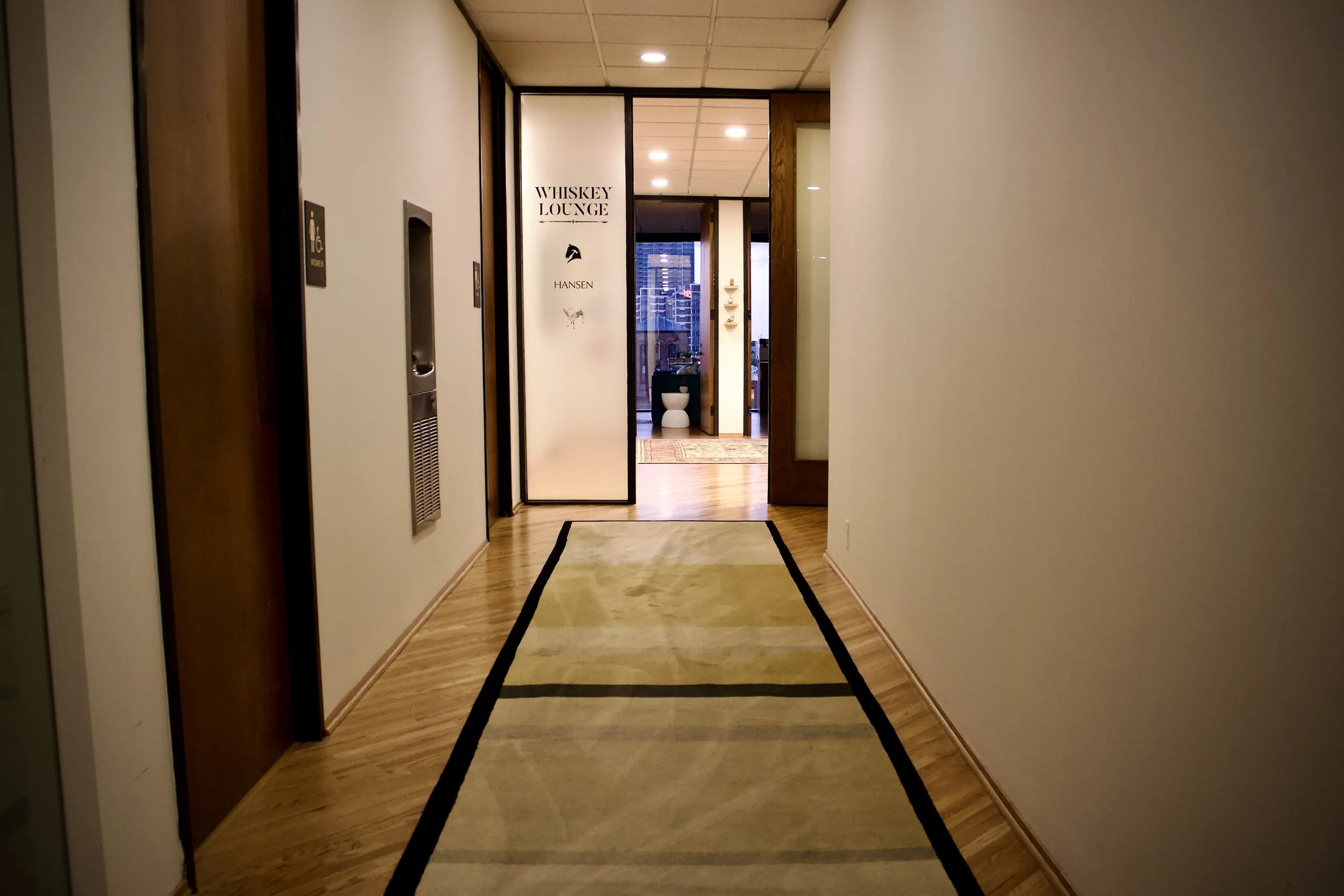 Hallway leading to the Whiskey Lounge in a building with wooden floors and a beige rug, signs indicating restroom and wheelchair accessible, with a window view of city buildings outside.