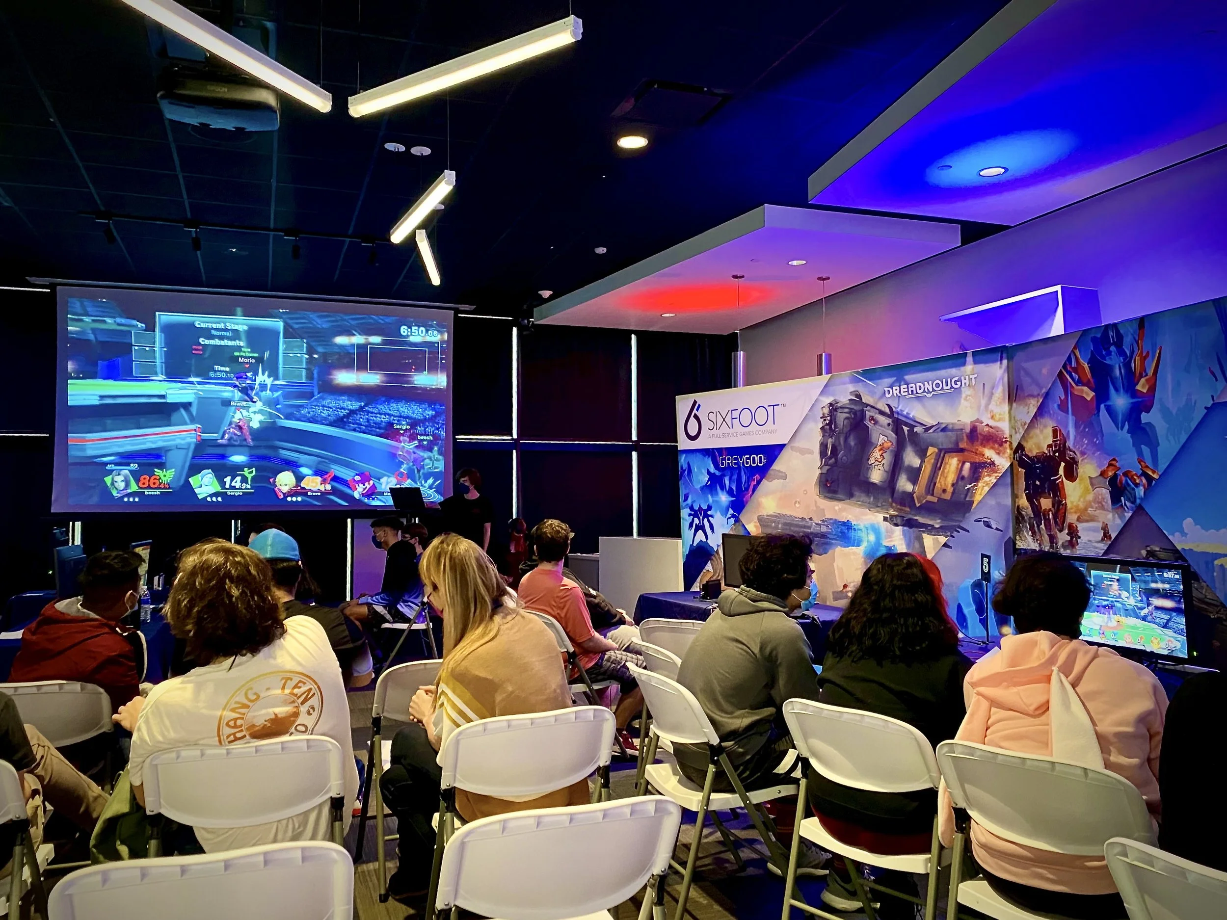 People sitting in chairs watching a video game tournament on a large screen in a dark room with colorful lights and promotional banners.