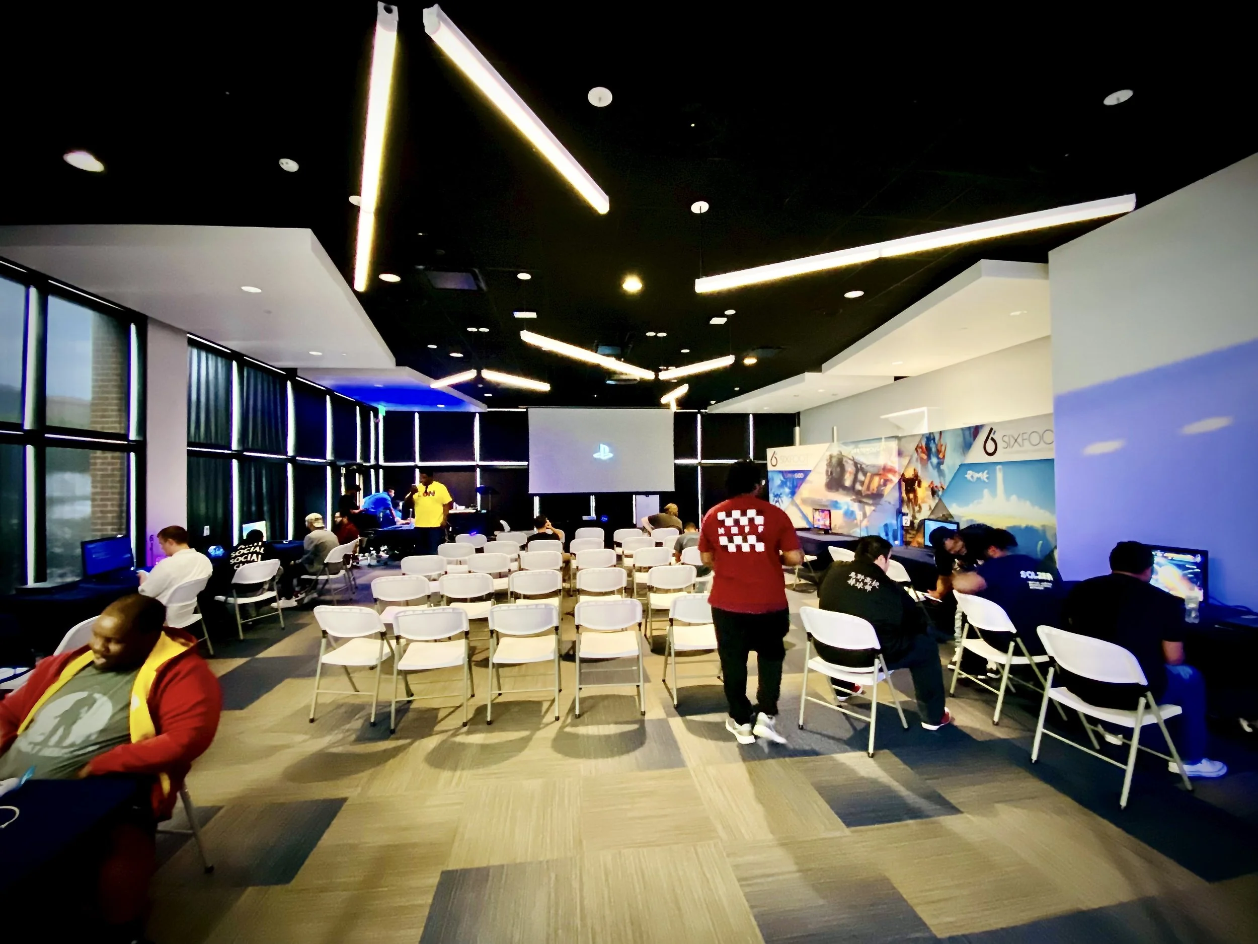 Indoor gaming event with attendees playing video games, some seated at computers, and a large screen at the front of the room.
