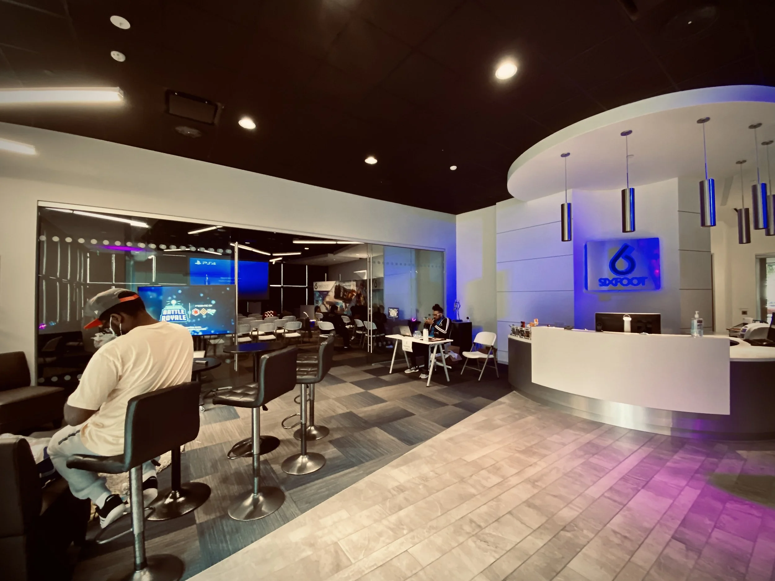 Interior of an internet cafe or gaming lounge with multiple people sitting at desks, large screens, and a reception area with a logo that says 'Swoot' on the wall.