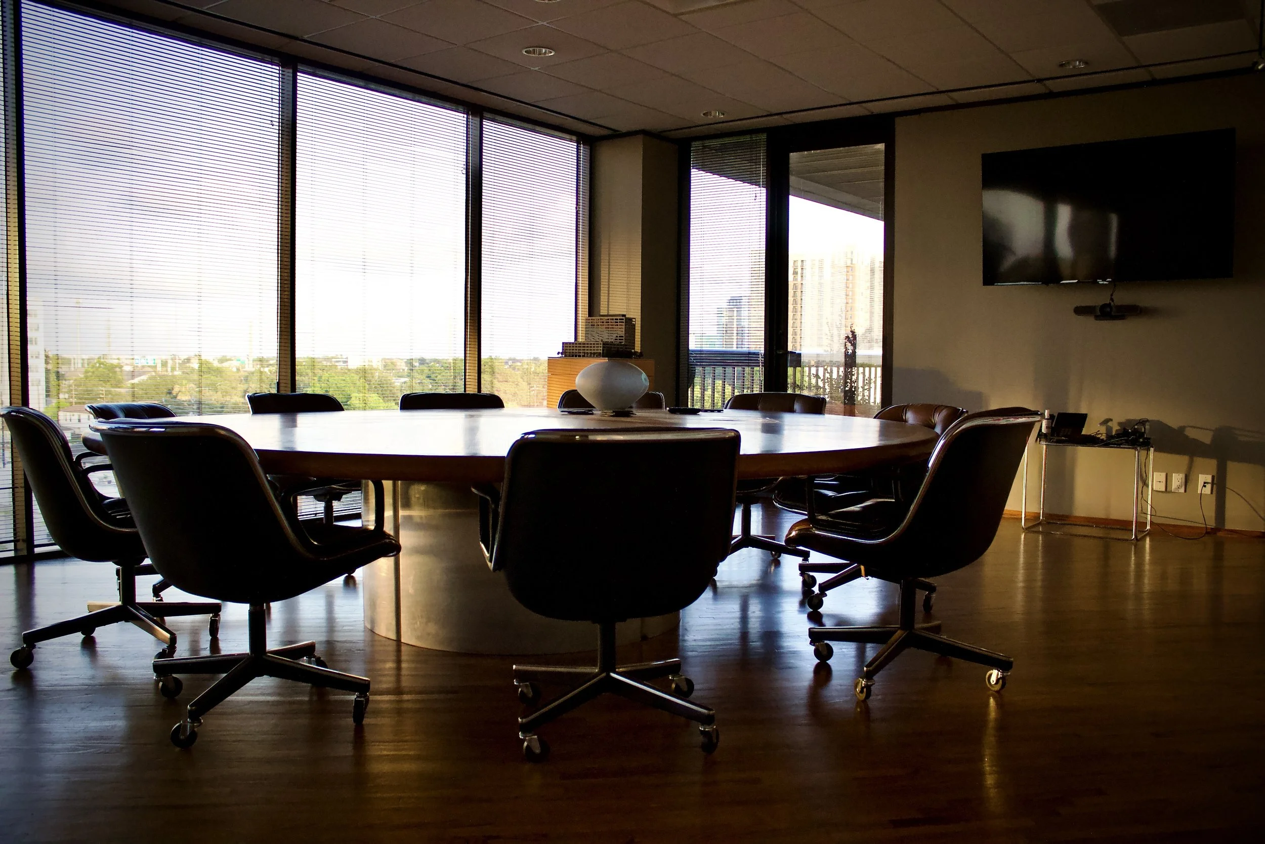 Empty conference room with a large round wooden table surrounded by black office chairs, large windows with blinds letting in natural light, a wall-mounted TV, and a small table with a bowl and office supplies.