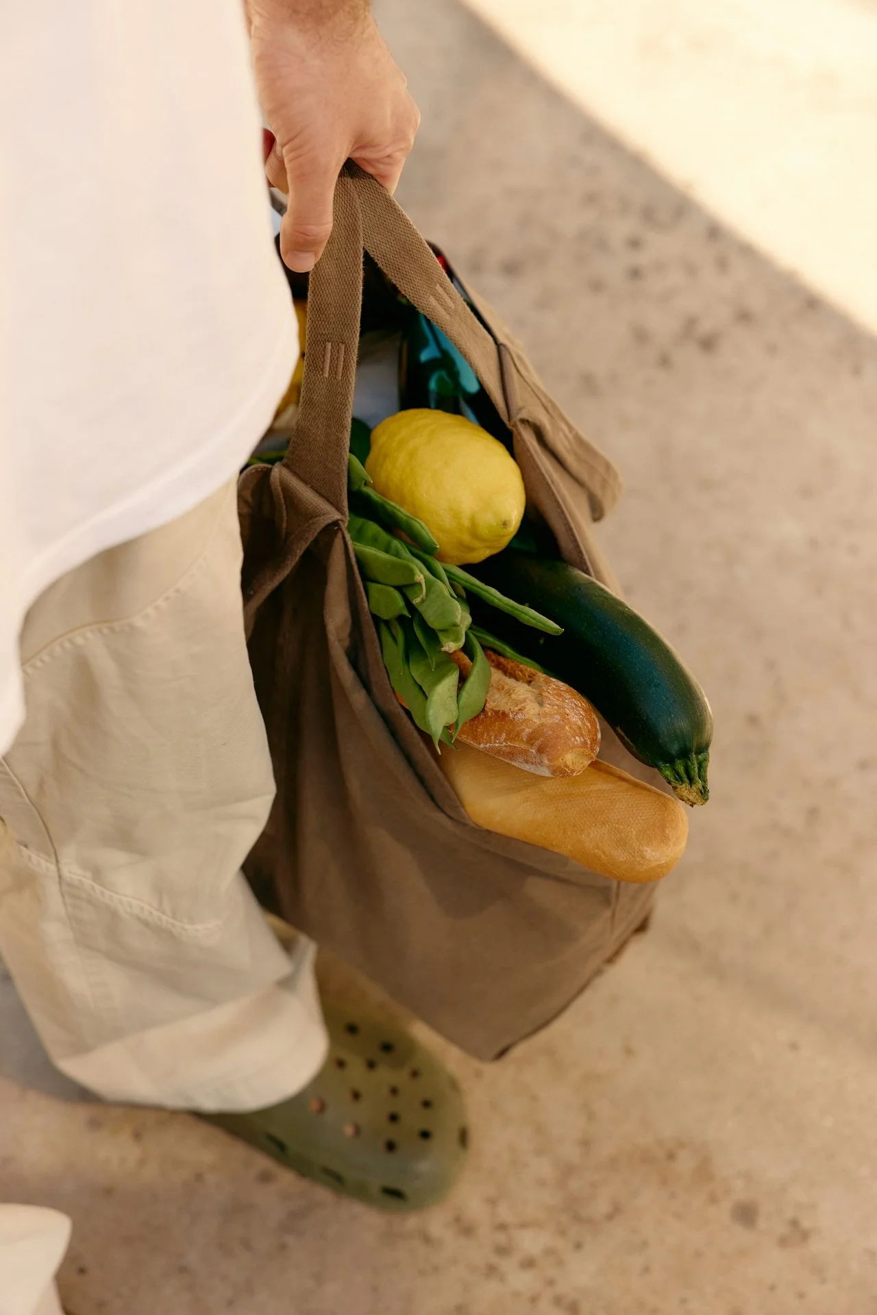 A person holding a brown reusable grocery bag filled with a lemon, green beans, a zucchini, and a baguette.