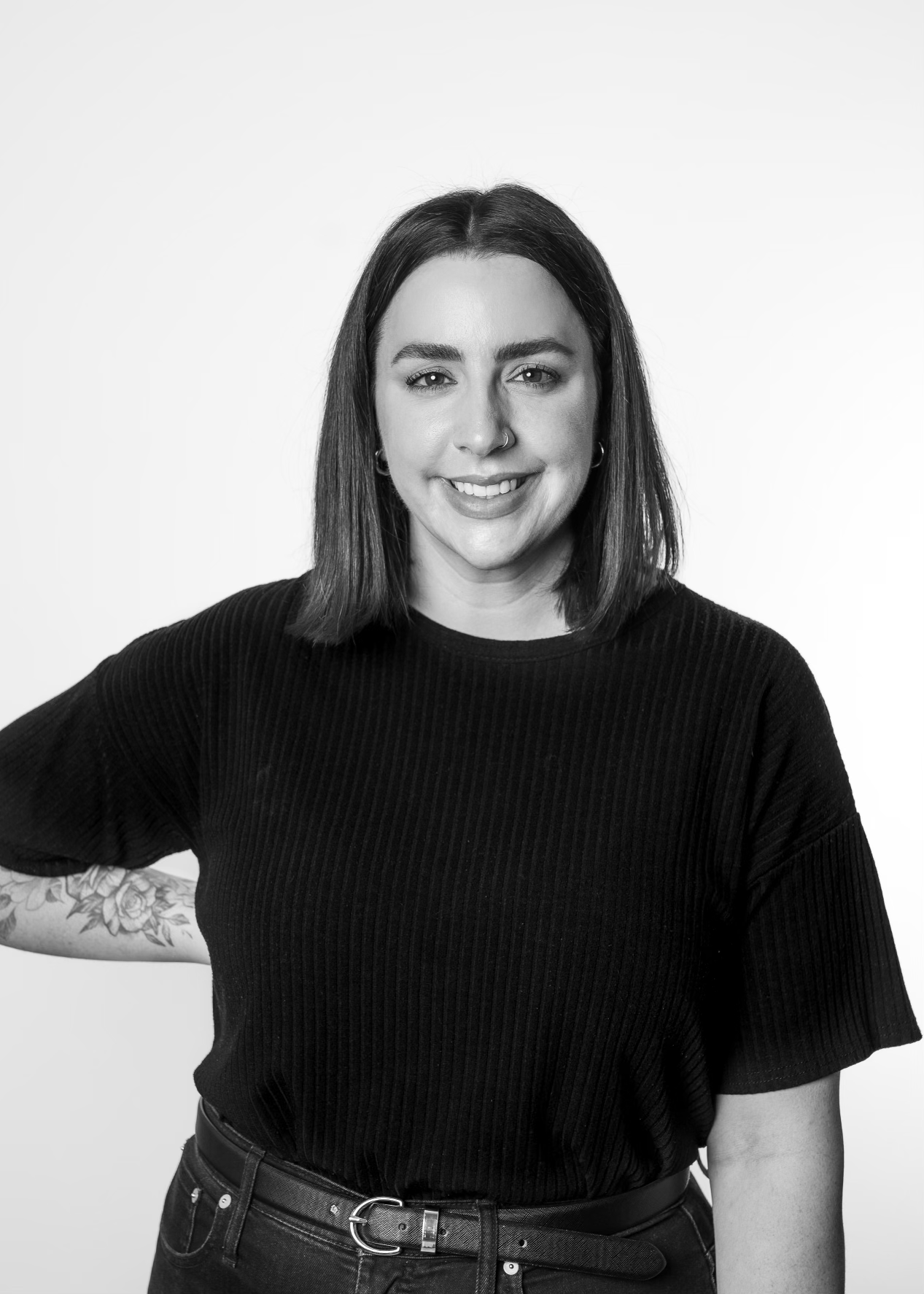 Black and white portrait of a smiling woman with shoulder-length dark hair, wearing a black ribbed top and a belt, standing against a plain background.
