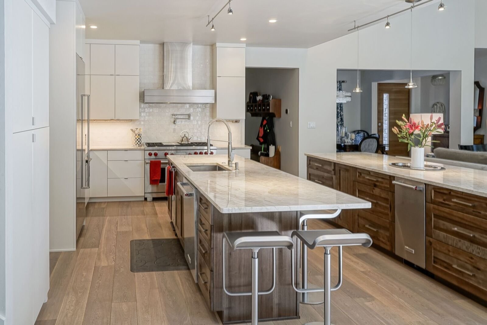 Modern kitchen with white cabinets, a large island with a marble countertop, wooden lower cabinets, stainless steel appliances, a flower vase on the island, and dining area visible in the background.