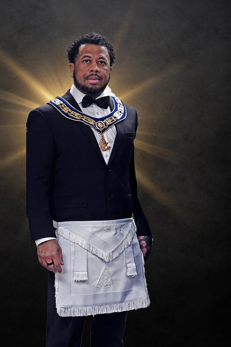 A man dressed in formal Masonic regalia, including a black tuxedo with a white apron, a bow tie, and a Masonic collar with gold details, standing against a dark background with radiant light effects.