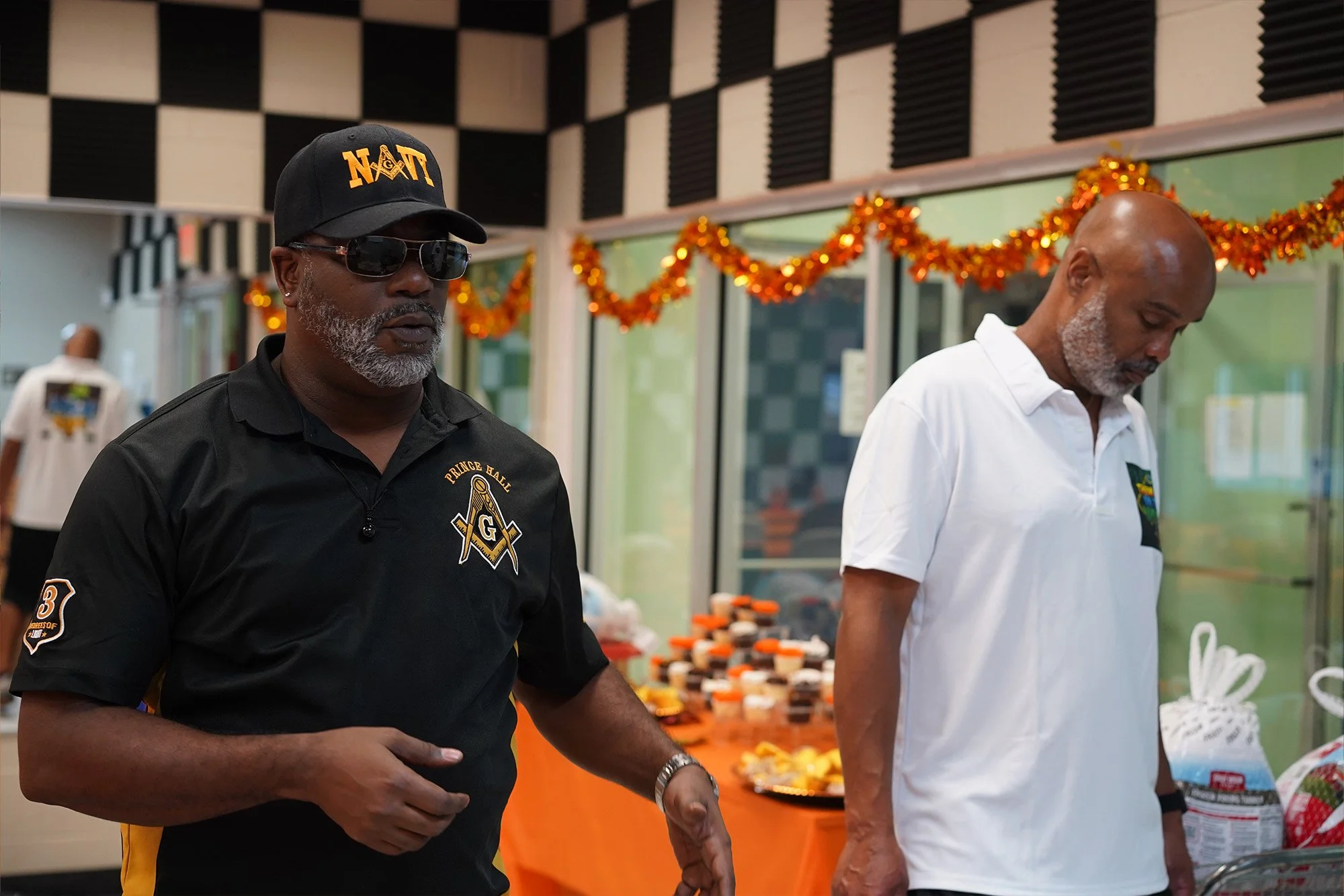 Two men with gray beards and black and white hair, respectively, are standing indoors, surrounded by Halloween decorations. The man on the left is wearing a black polo shirt with Masonic symbols, a cap with 'NAT' embroidered on it, and sunglasses. The man on the right is wearing a white shirt and is looking down. There are Halloween orange and black decorations hanging on the wall in the background and a table with Halloween treats.