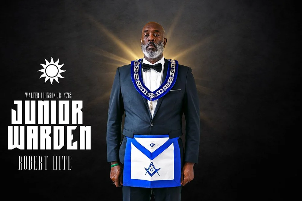 A man in a suit wearing a Masonic apron and a blue Masonic collar, standing in front of a dark background with a gold halo-like glow, with the text "Junior Warden" and "Robert Hite" on the left side, and a white sun symbol above the text.