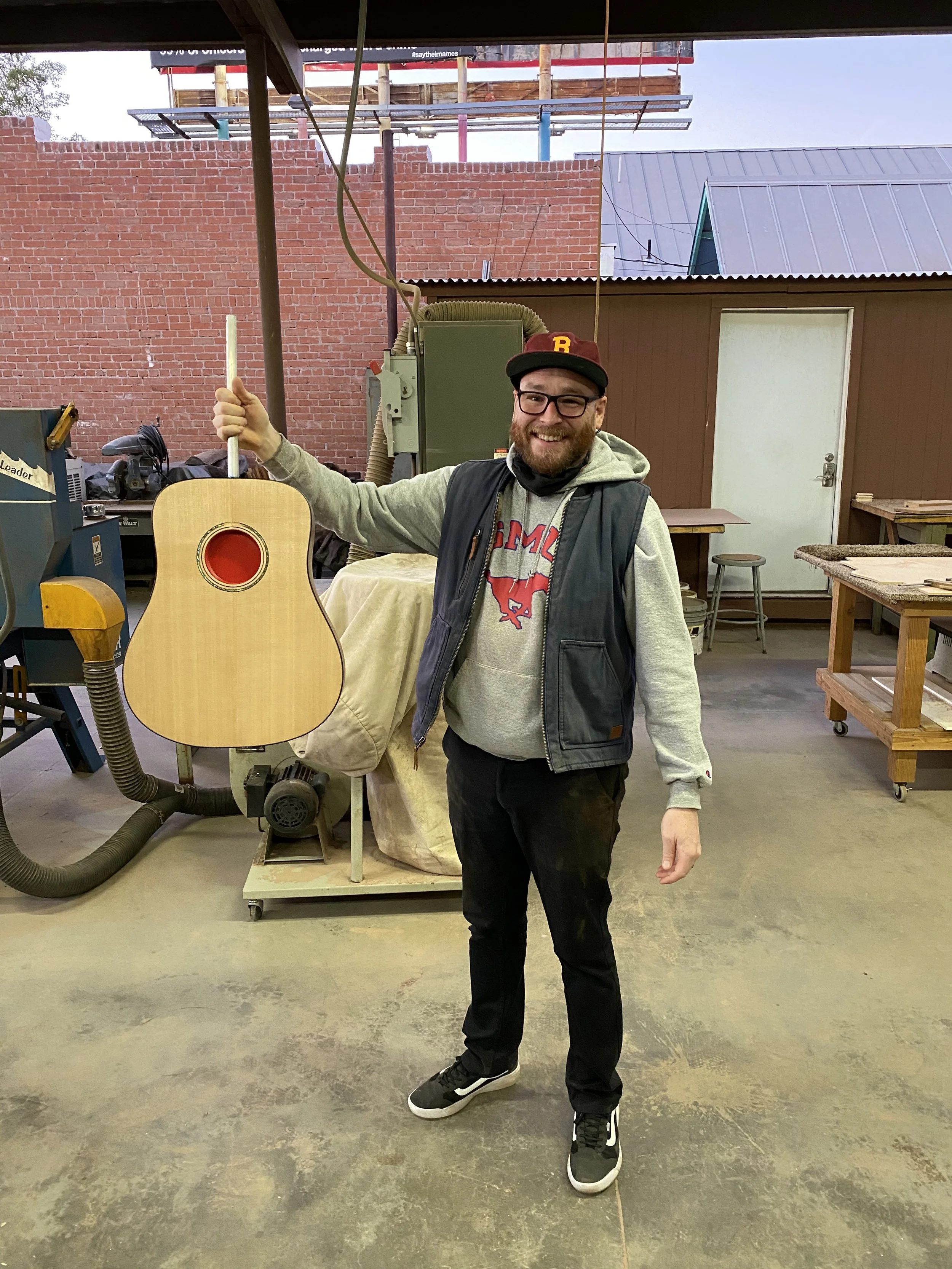 A man smiling and standing in a woodworking shop, holding an unfinished acoustic guitar next to a workbench and woodworking equipment.