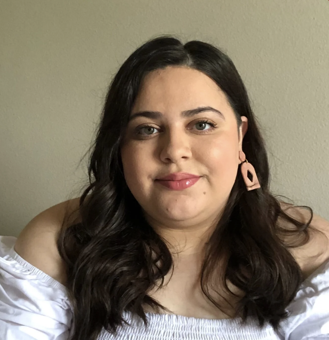 A woman with dark hair, wearing pink earrings and a white off-the-shoulder top, smiling in front of a beige wall.