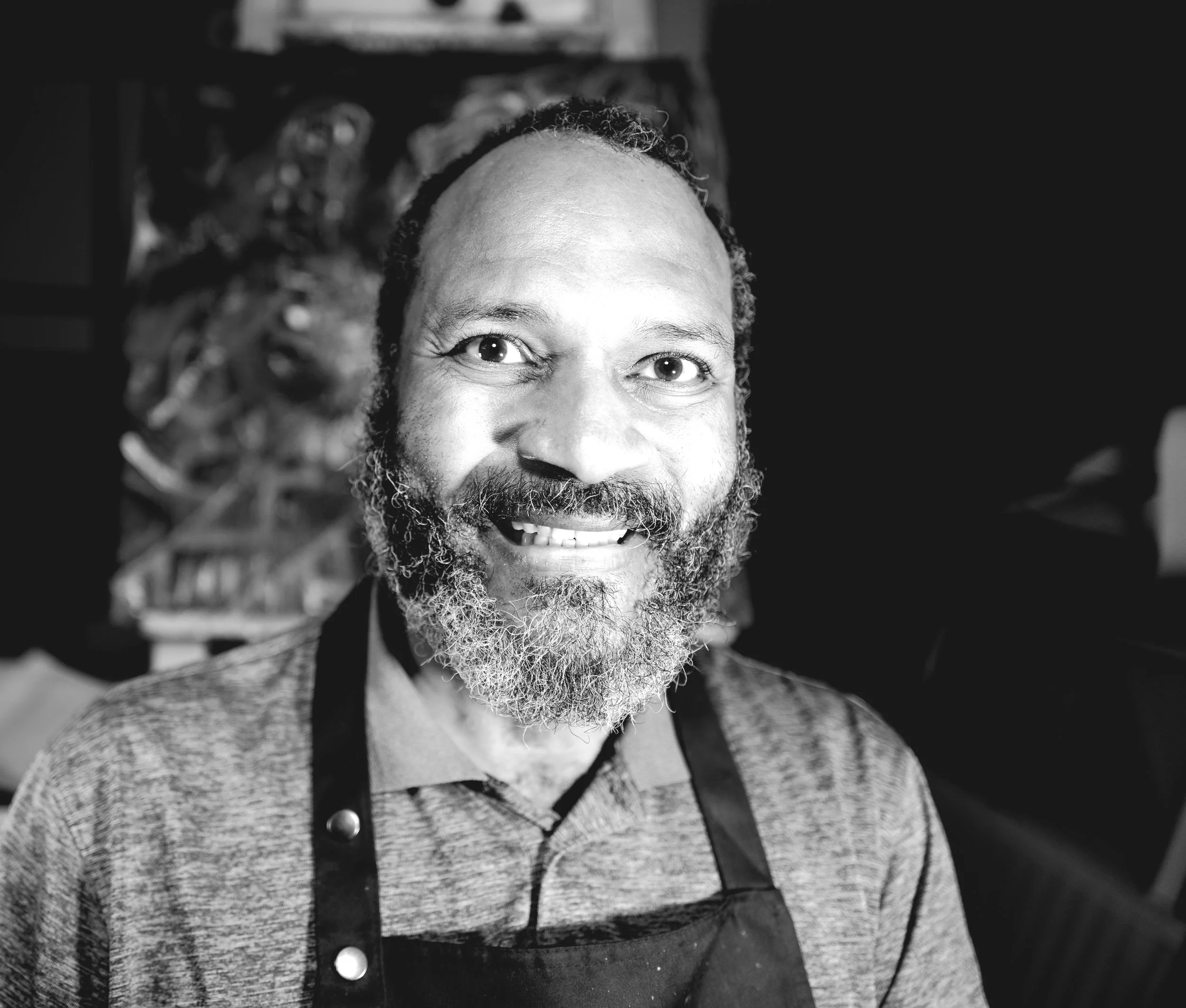 A smiling man with a beard and dreadlocks wearing an apron in black and white. Background features a blurry painting or artwork.