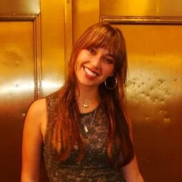 Young woman with long brown hair smiling, wearing hoop earrings and a sleeveless dress in a warmly lit room.