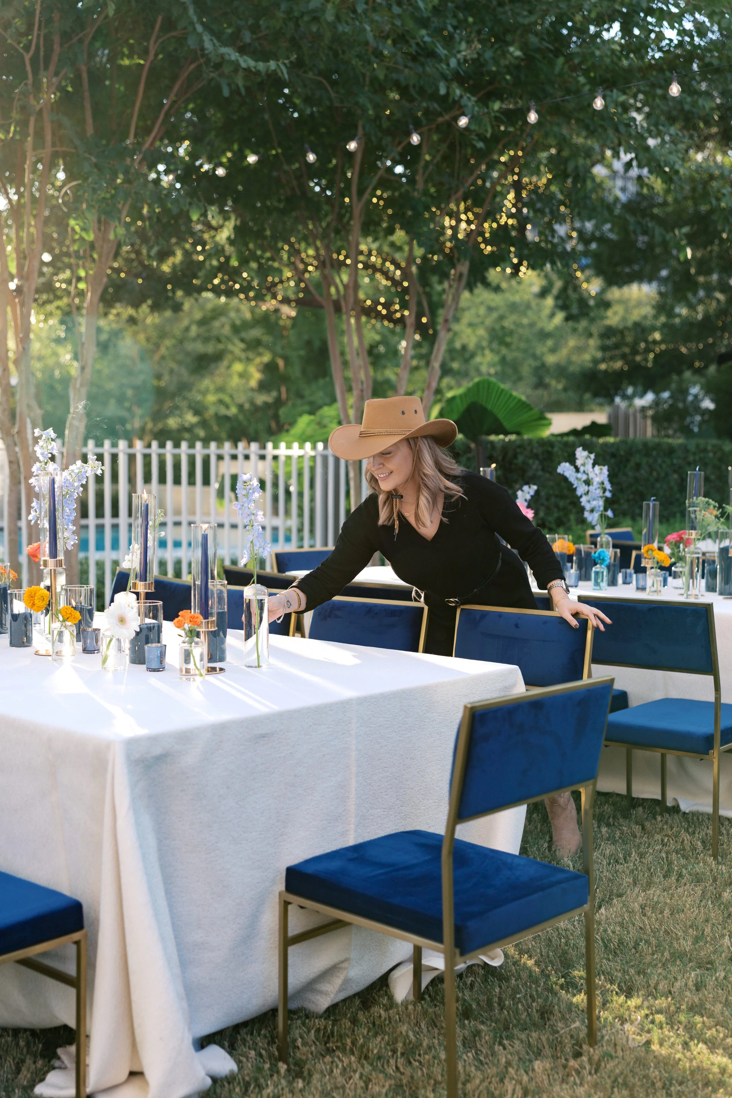 Woman arranging table decorations at an outdoor event in a garden setting with string lights and trees.