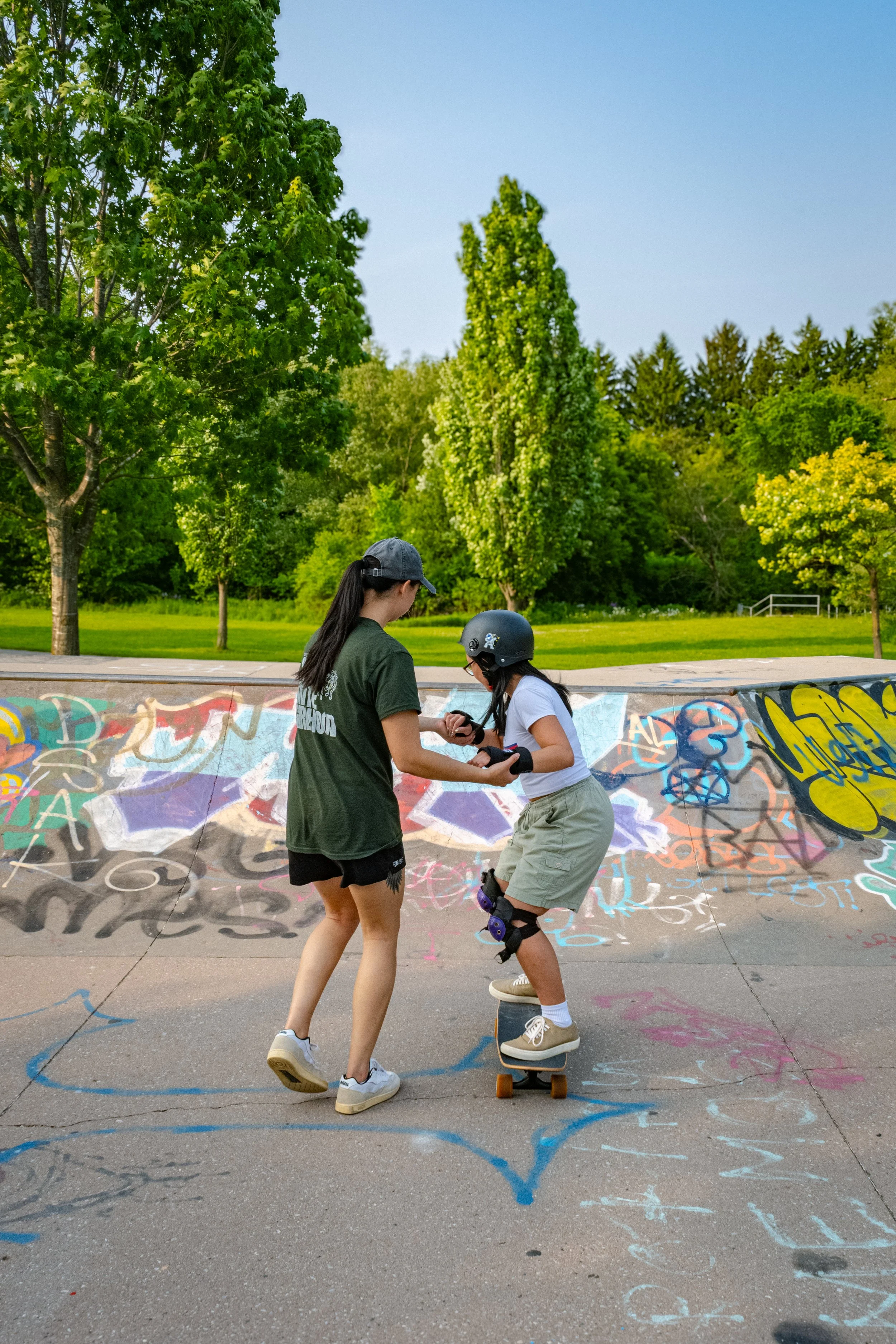 Two girls at a skate park, one assisting the other with her skateboard. The girl on the skateboard is wearing a helmet, knee pads, and casual clothes. The skate park has graffiti on the inclined surface, and trees and a grassy area are visible in the background.