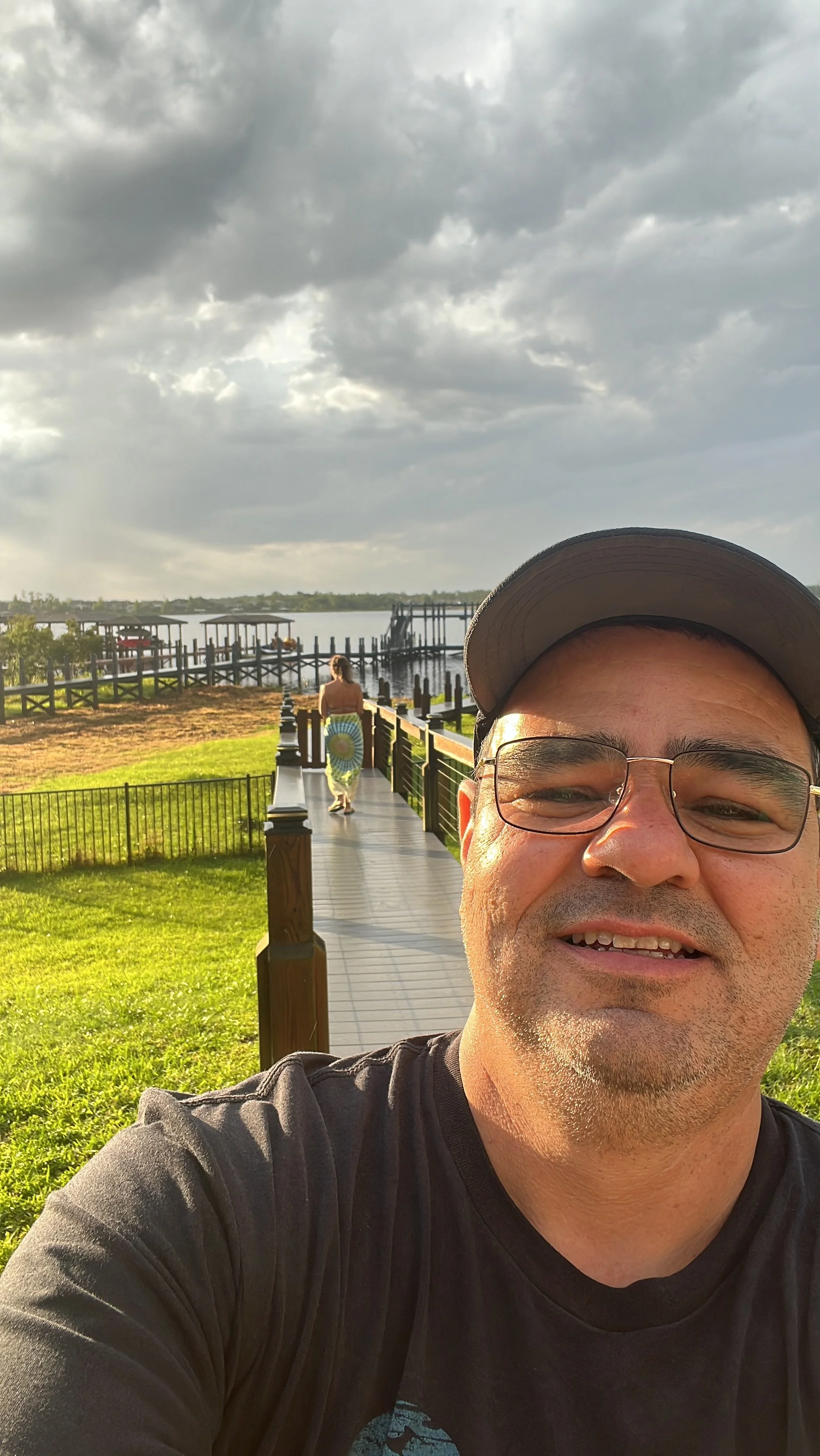 Self-portrait of a man wearing glasses and a cap taking a selfie outdoors near a river with a dock, a woman walking away in the background, green grass, and a cloudy sky.