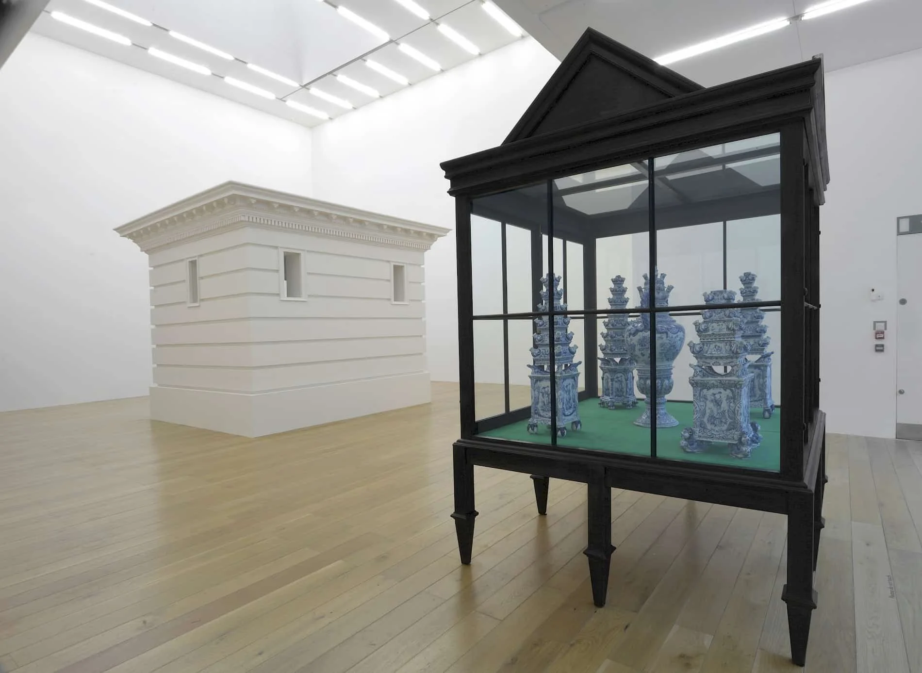 Art exhibit with a white building model and an enclosed display case with blue and white porcelain vases and sculptures in a gallery with wooden floors and white walls.