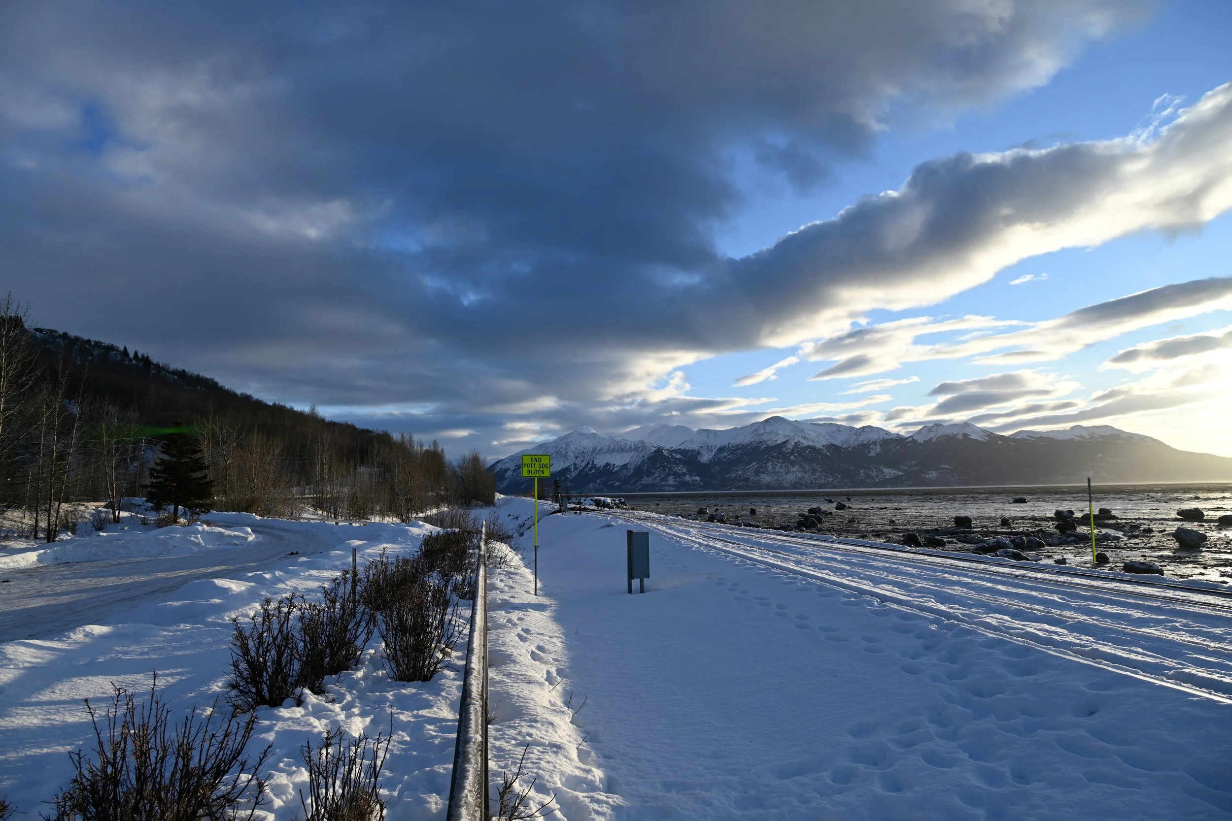 Snowy road and landscape with mountains in the distance, cloudy sky, and a yellow sign that reads 'End Pott SDC Block'.