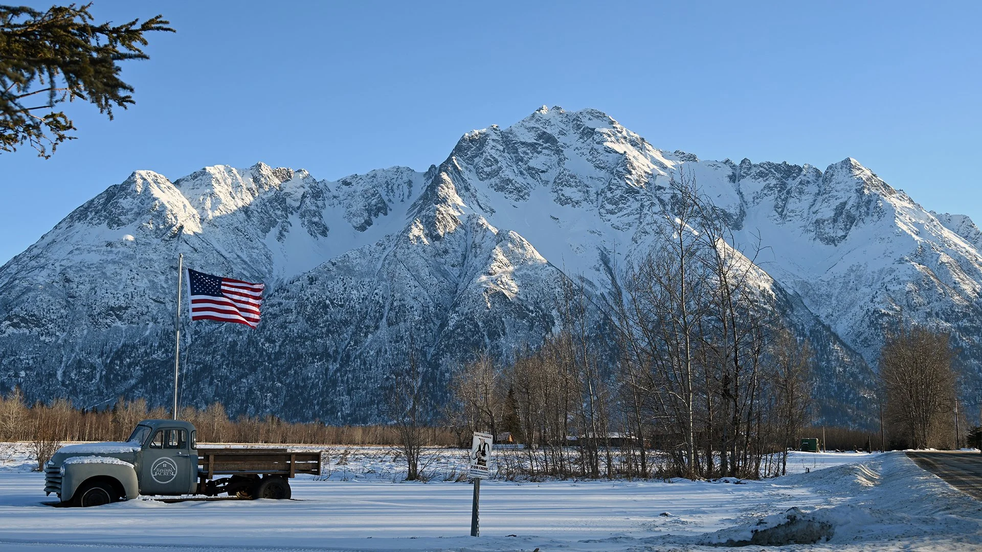 A snow-covered landscape with a vintage truck, an American flag, and tall mountains in the background.