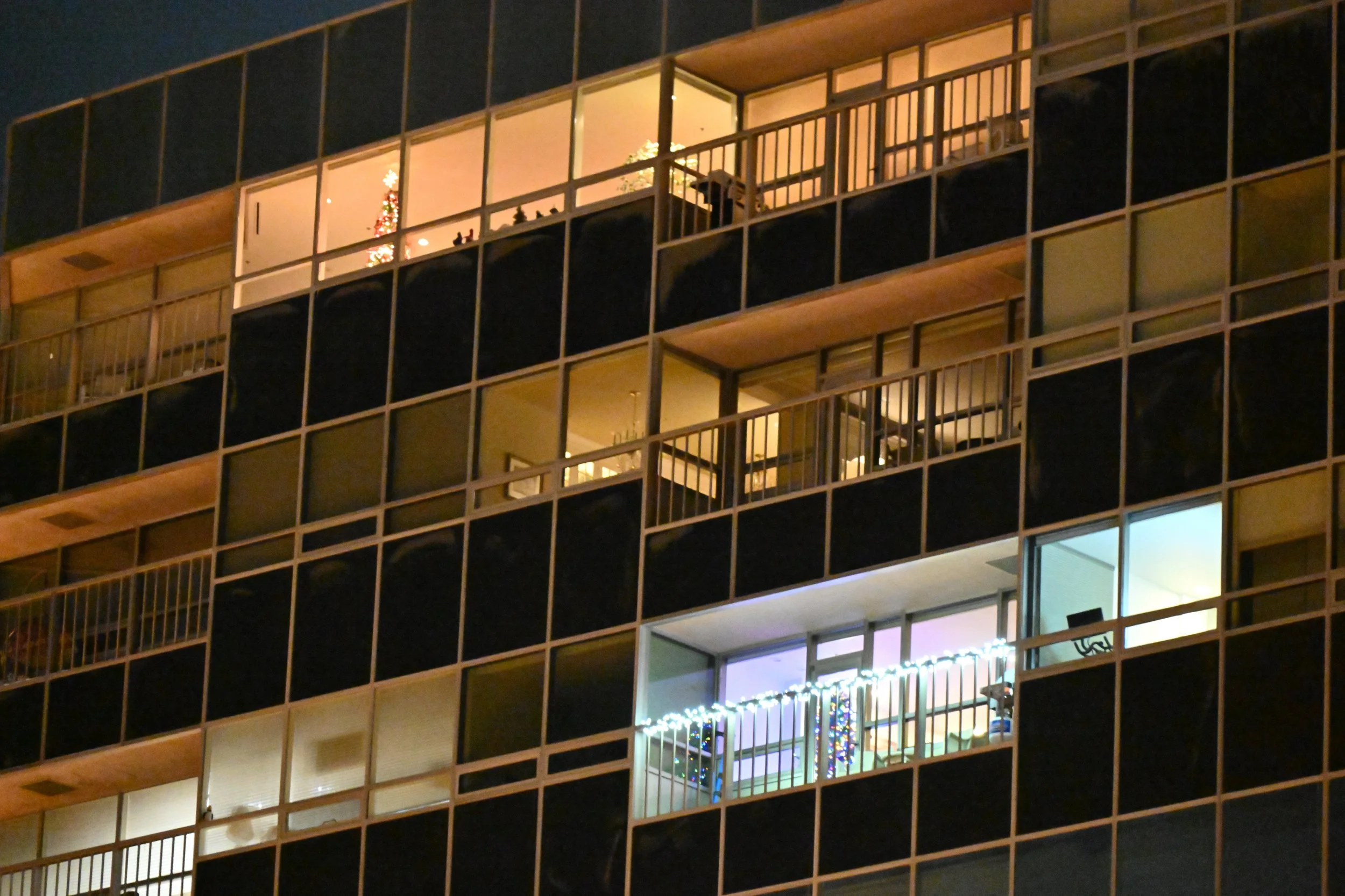 Nighttime view of a multi-story apartment building with lit windows and decorated Christmas lights on one balcony.