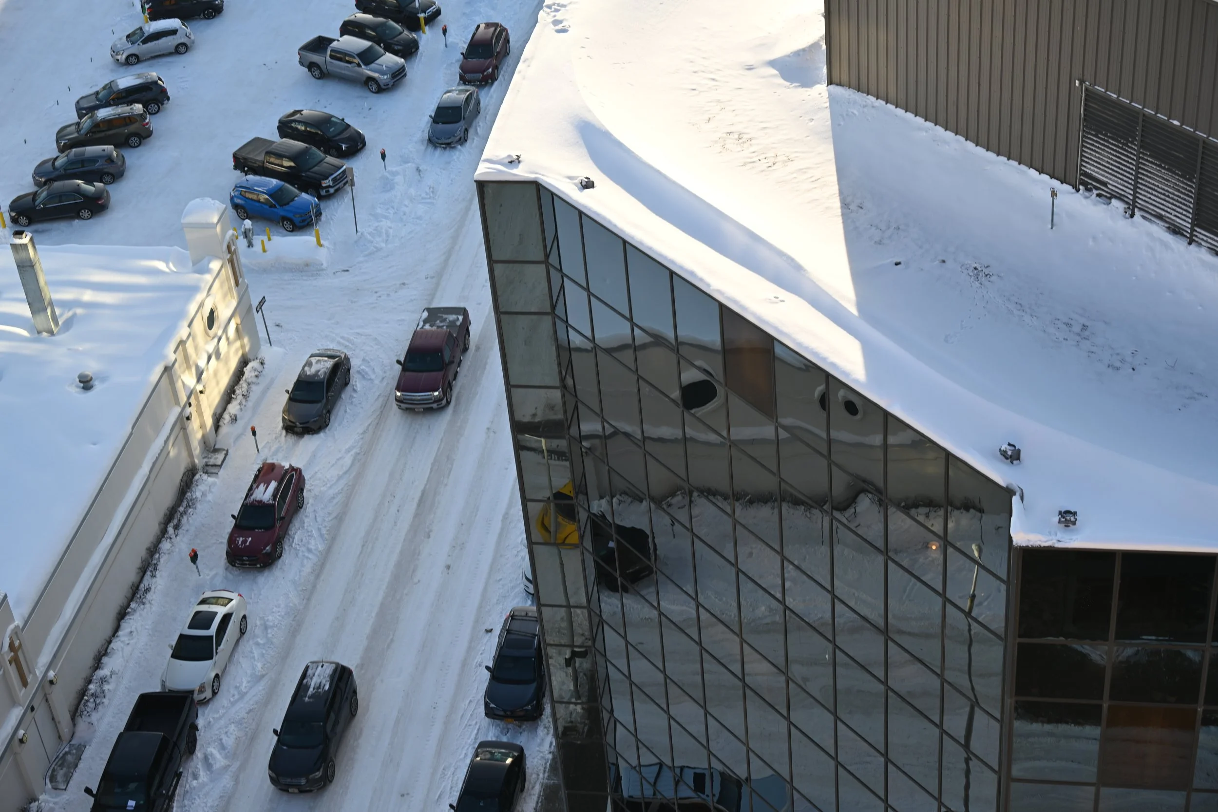 Aerial view of a snowy city street with parked cars and a modern glass building reflecting the surrounding environment.