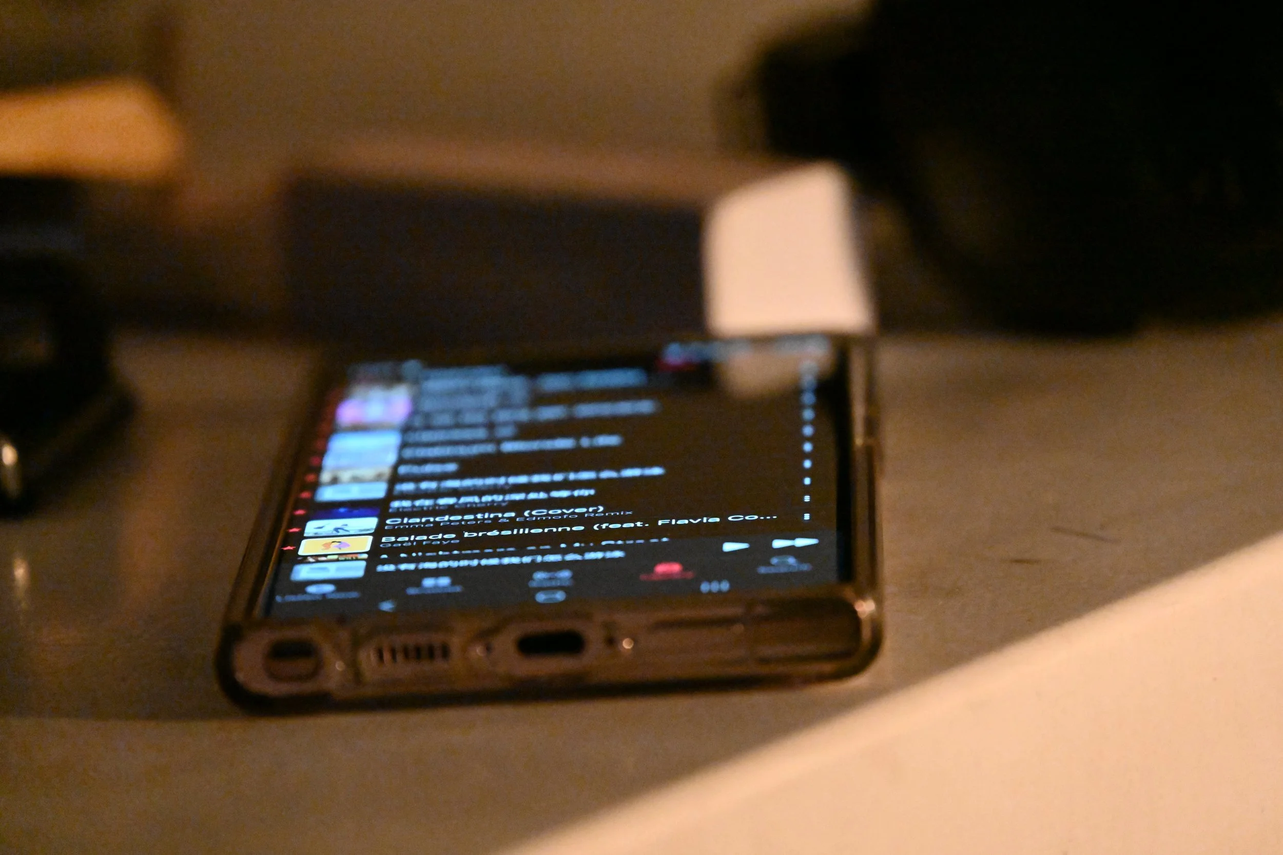 A smartphone lying on a table, displaying a music playlist in dark mode with colorful album art and text, with a white lamp and a blurred background.