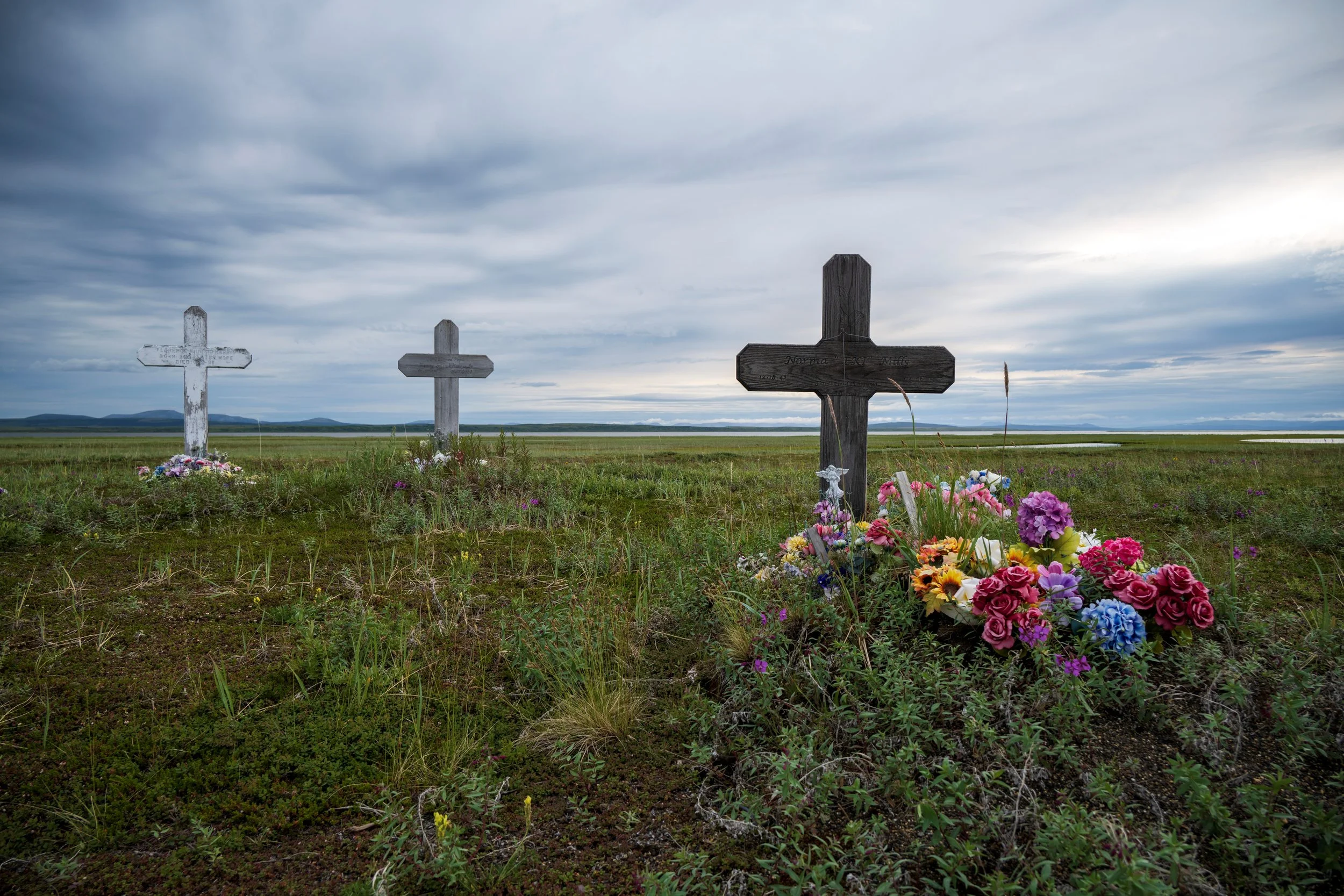 Three graves with crosses in a grassy field under cloudy sky, one decorated with colorful flowers.