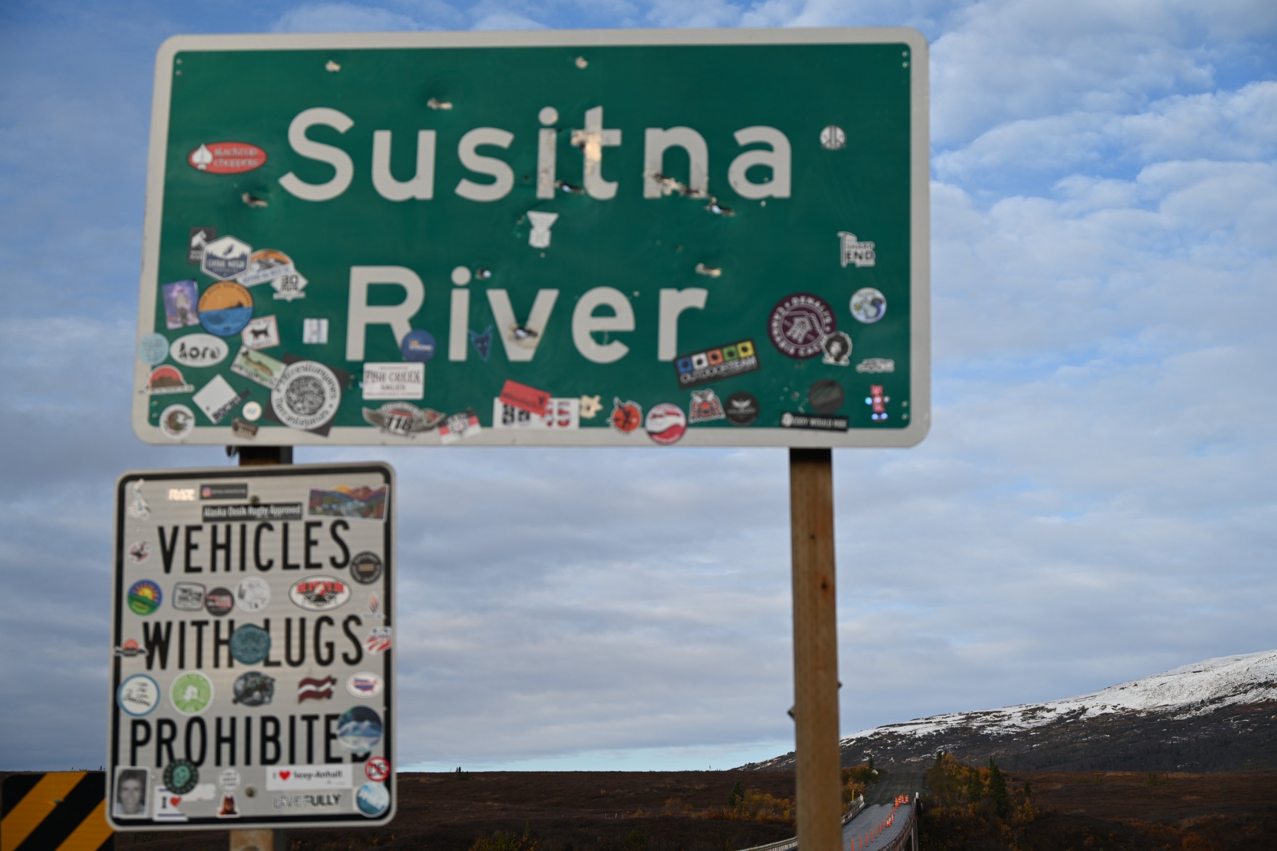 A green highway sign reading "Susitna River" with many stickers on it, and a black and white sign underneath that says "Vehicles with Lugs Prohibited" with stickers. The background shows a cloudy sky, mountains, and a bridge on a rural road.