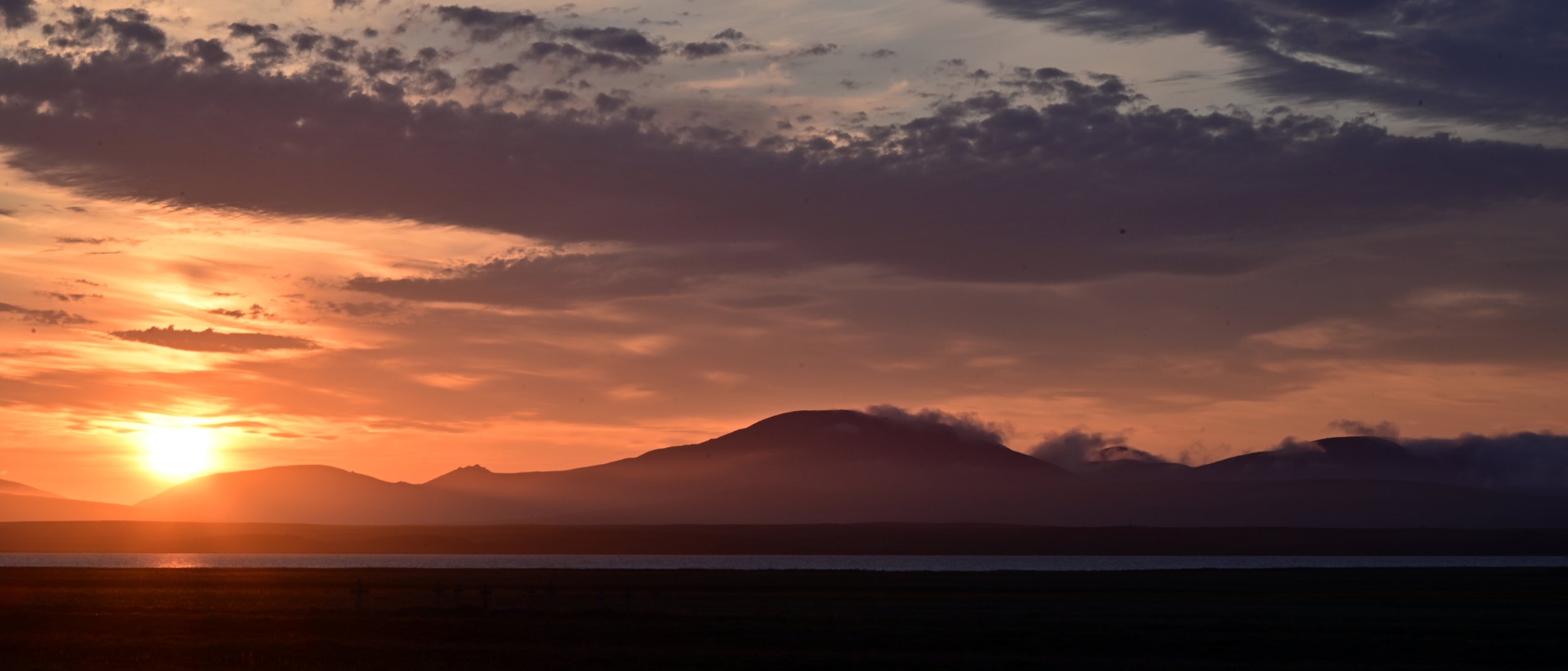 Sunset over a mountain range with clouds in the sky and a body of water in the foreground.