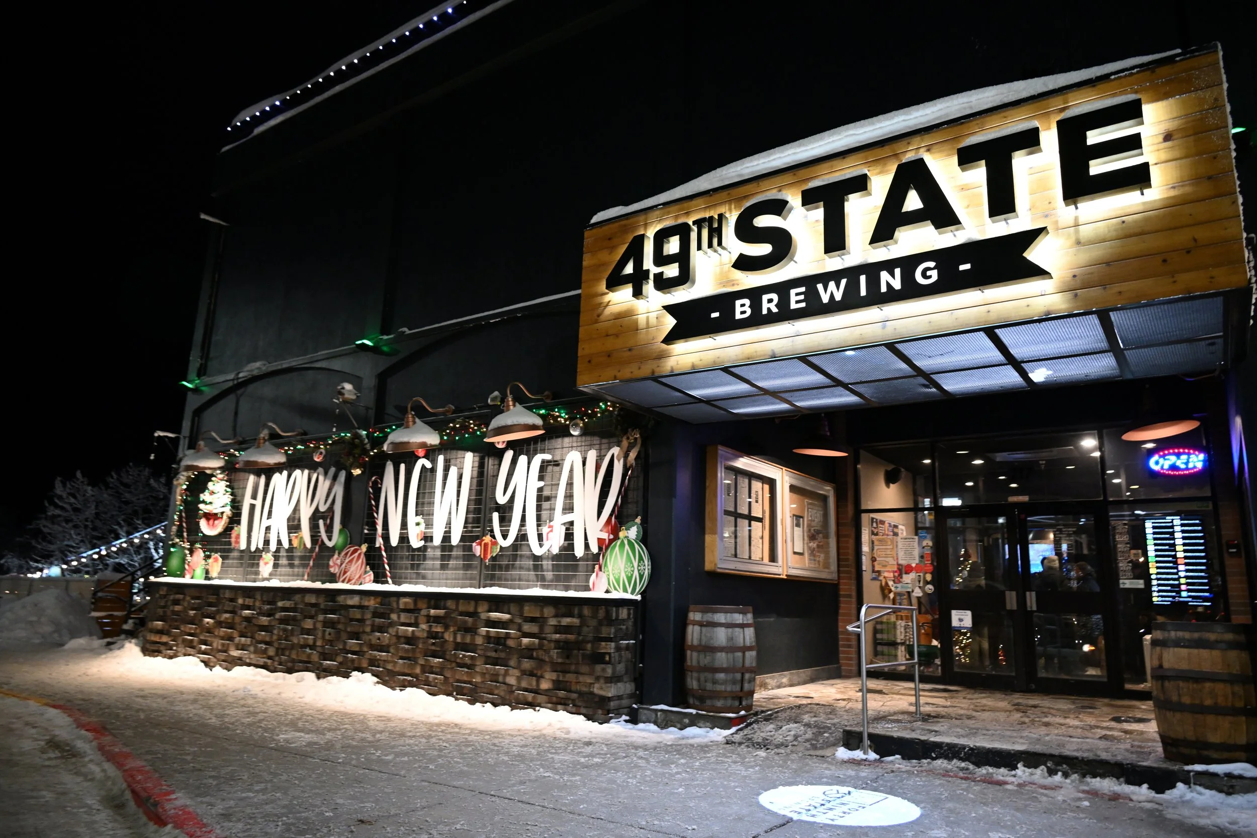 Night view of 49th State Brewing with holiday decorations, including a banner reading 'Happy New Year,' snow on the ground, and a lit 'Open' sign in the entrance.