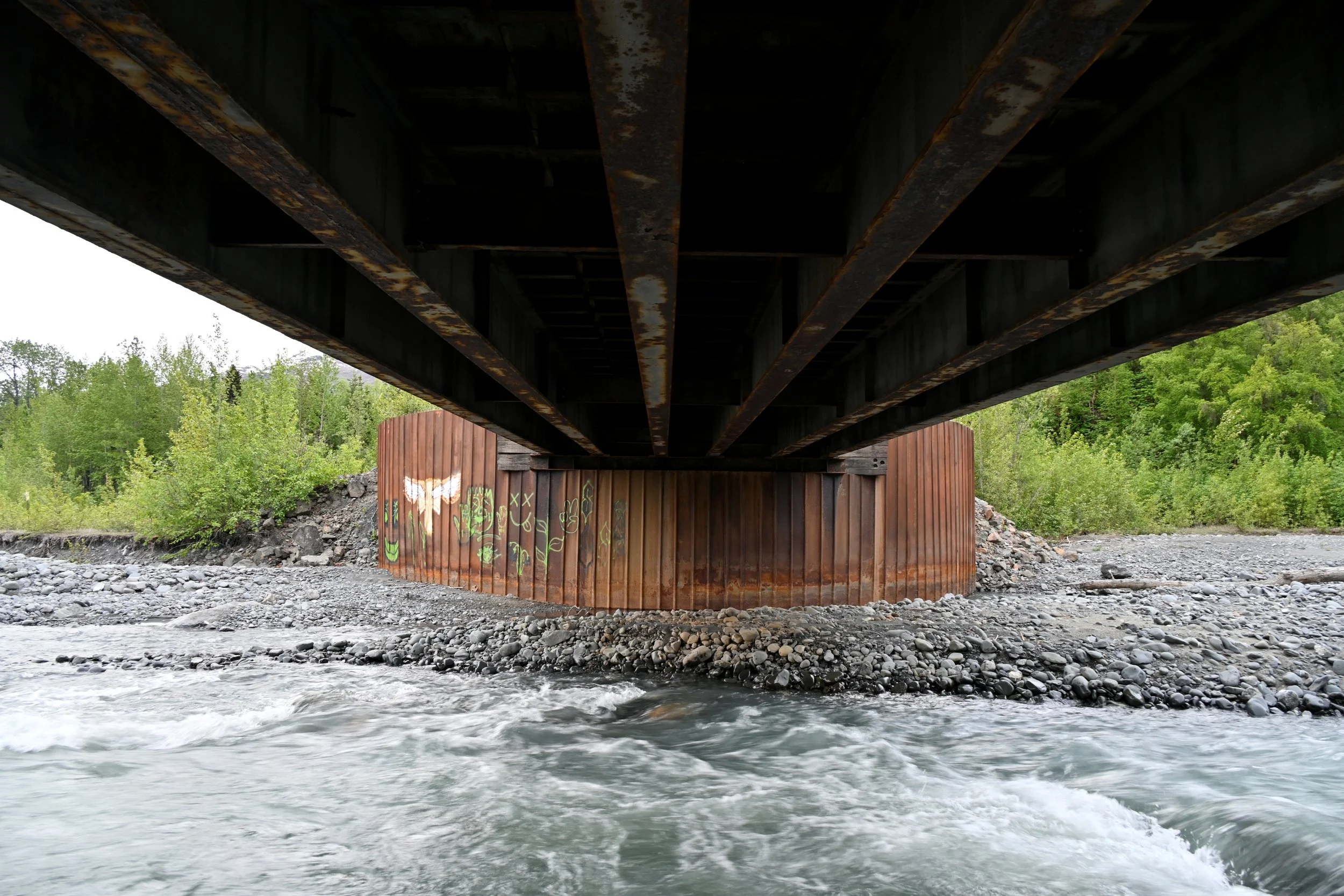 Underneath view of a bridge over a river, showing rust-streaked, circular wooden supports and graffiti with an owl and plants, surrounded by rocks and green trees.