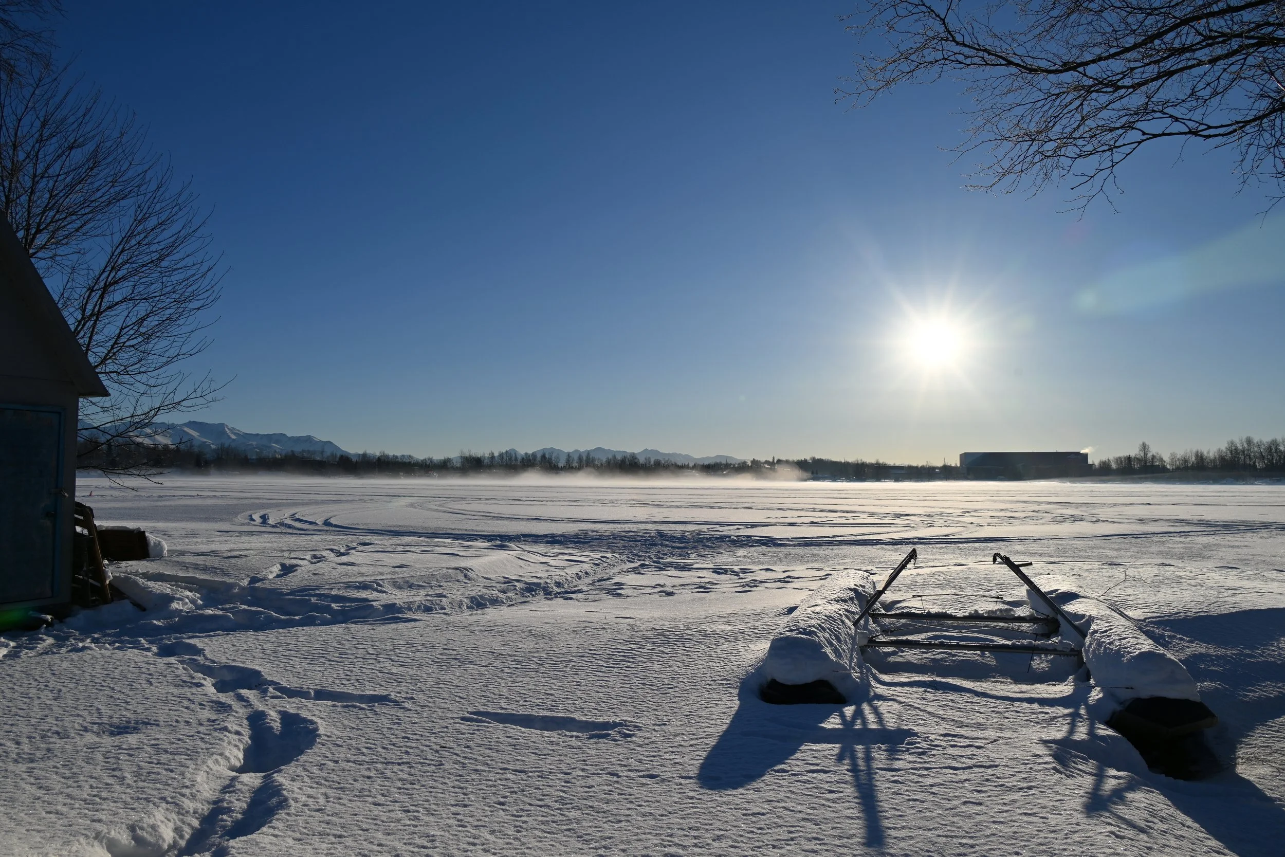 Snow-covered landscape with a bright sun in the sky, bare trees, and mountains in the distance. A sled lies on the snow in the foreground.