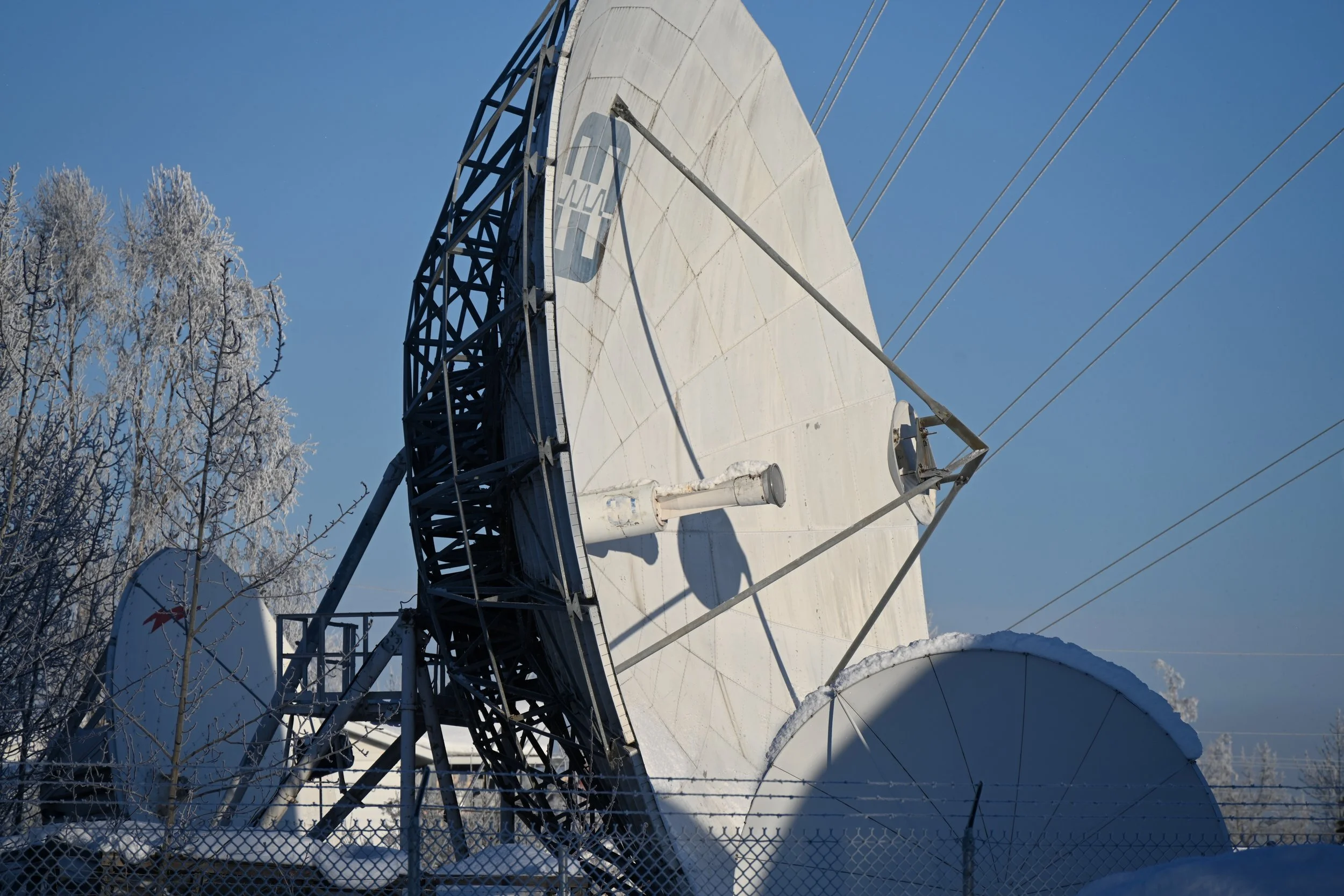 Large radio telescope dish covered in snow, with a black metal framework and a fence in the foreground, blue sky in the background, and snow-covered trees.