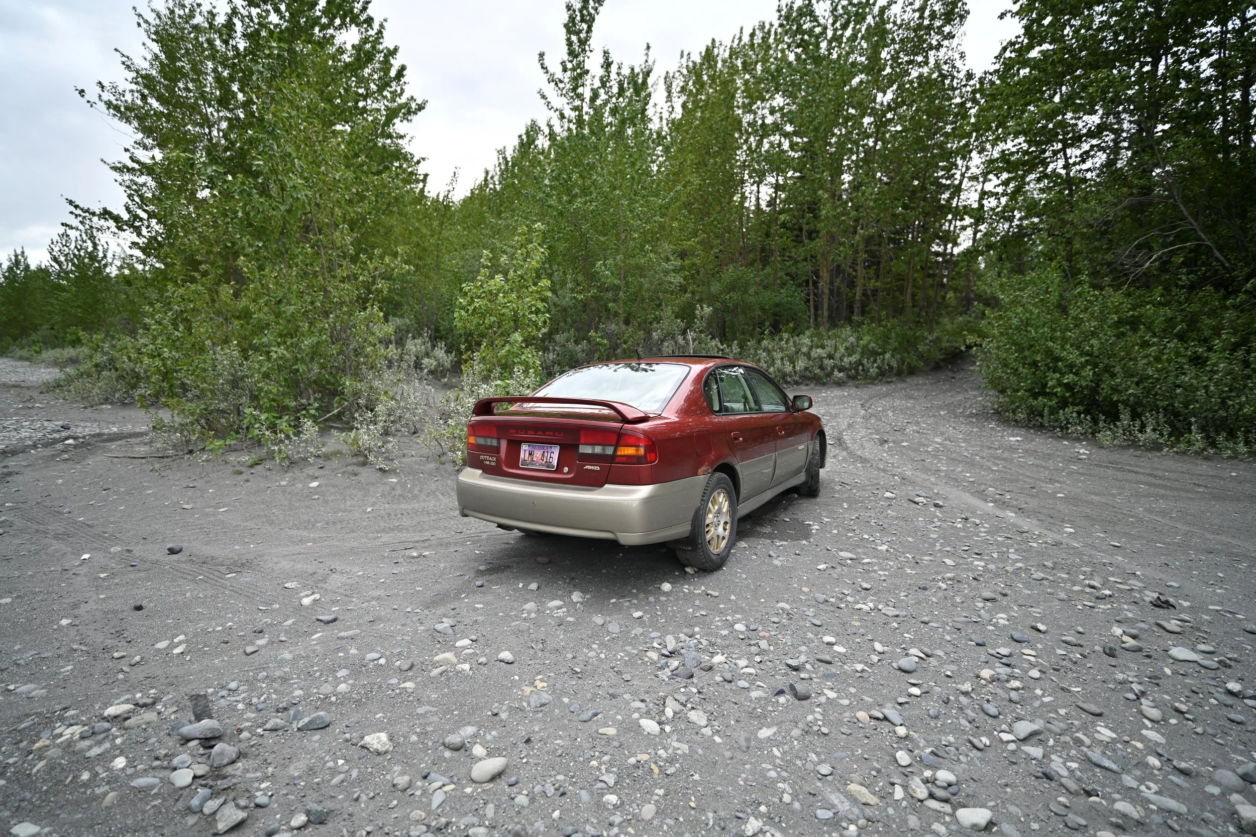 Red and beige sedan car parked on a gravel road near a forested area with trees and green foliage.