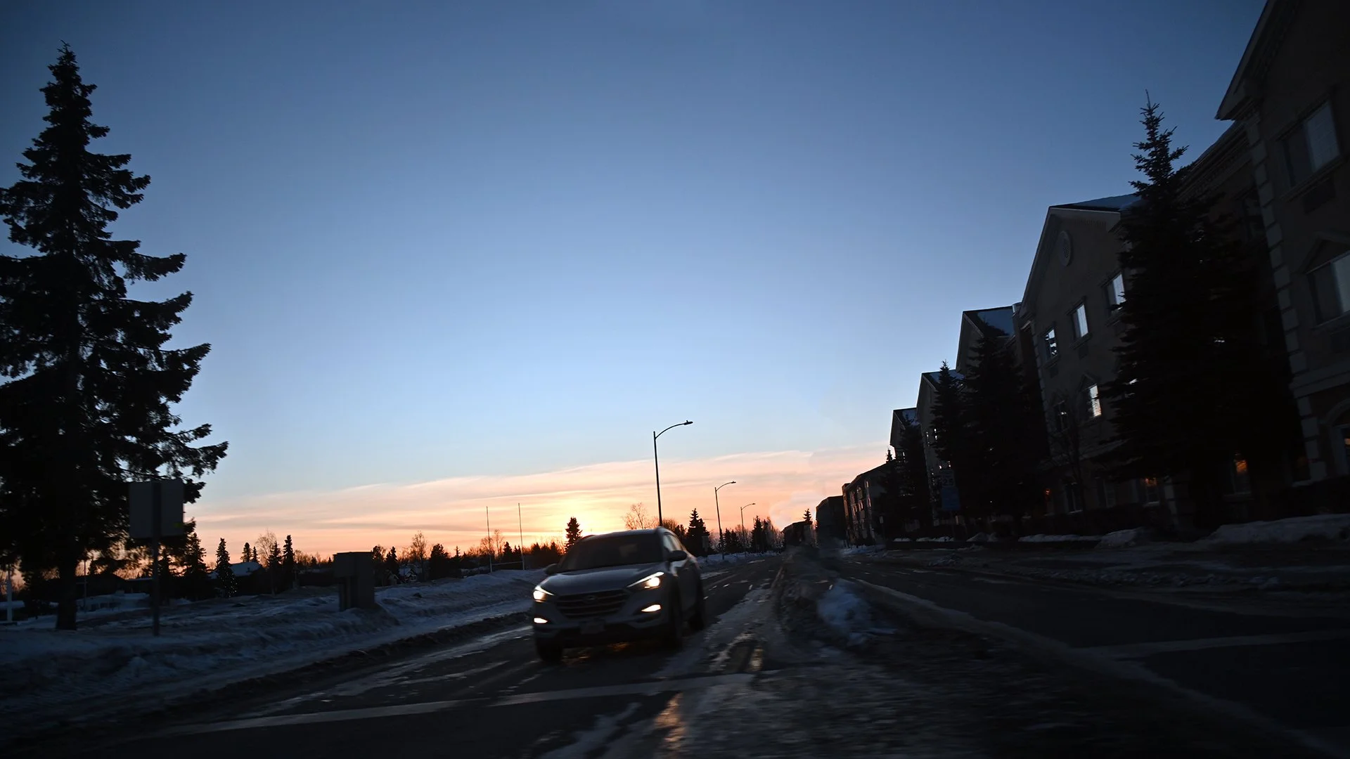A car driving on a residential street at dusk, with snow on the ground and a sunset on the horizon.